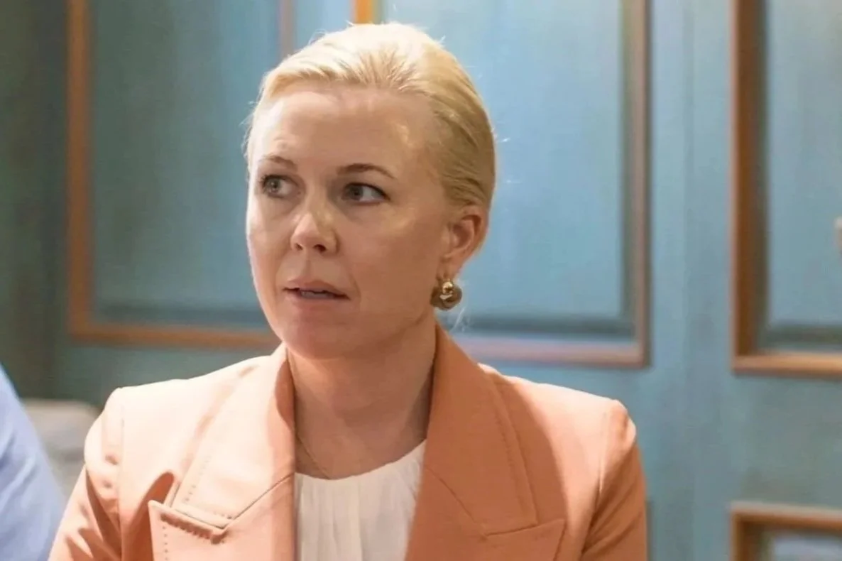 A middle-aged woman with blonde hair, wearing a peach blazer and gold earrings, sitting in an indoor setting with blue walls and framed artwork in the background.