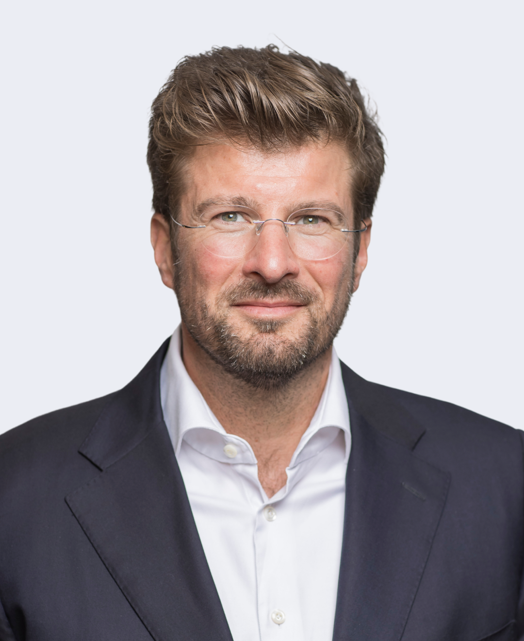 A middle-aged man with brown hair, glasses, and a beard, wearing a black suit jacket and a white dress shirt, smiling slightly against a plain light background.
