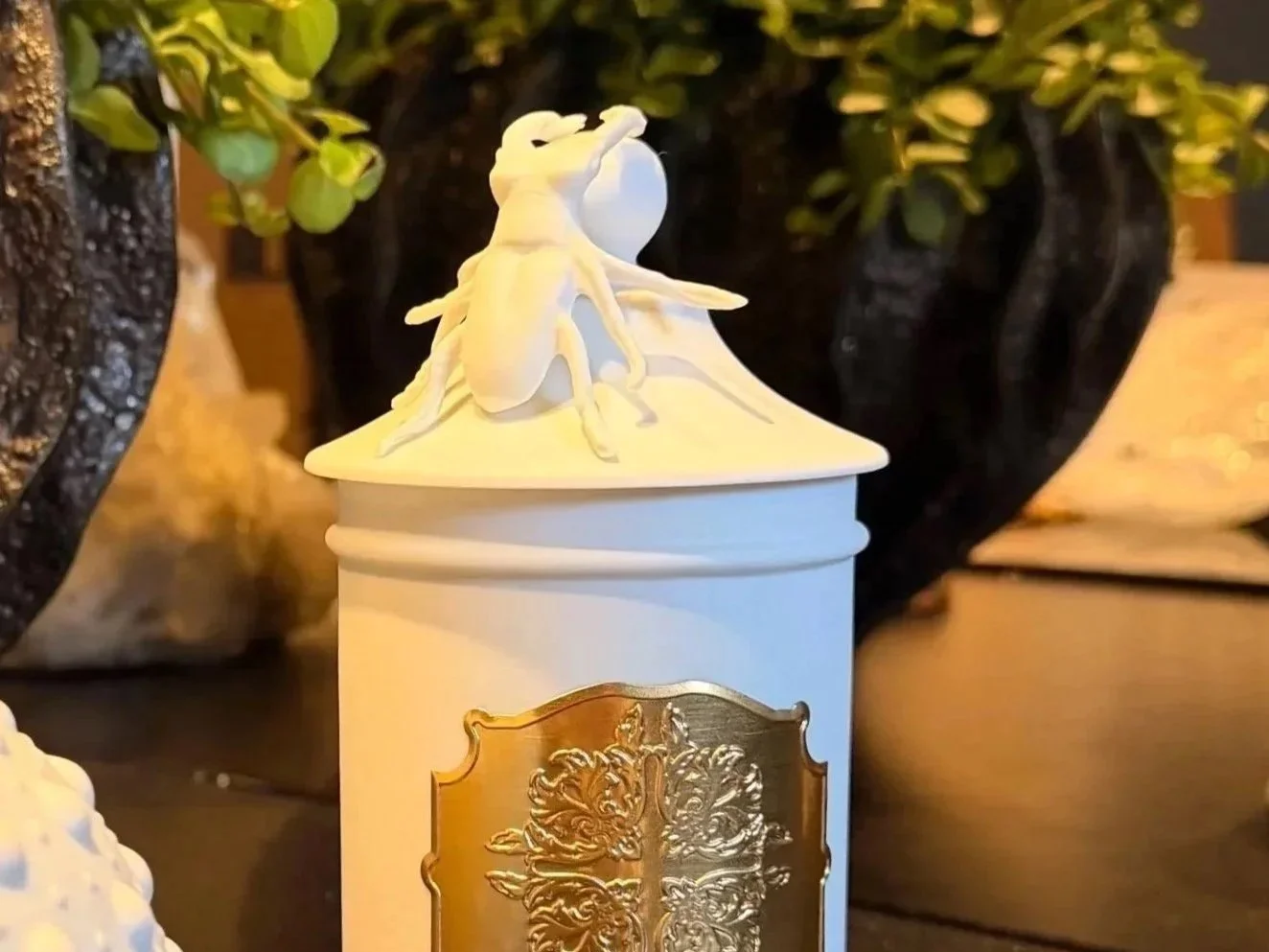 Une boîte blanche avec un haut en forme de toit, décorée d’un hamster en train de manger ou boire, posée devant un grand pot brun contenant une plante verte. La scène est éclairée chaleureusement.