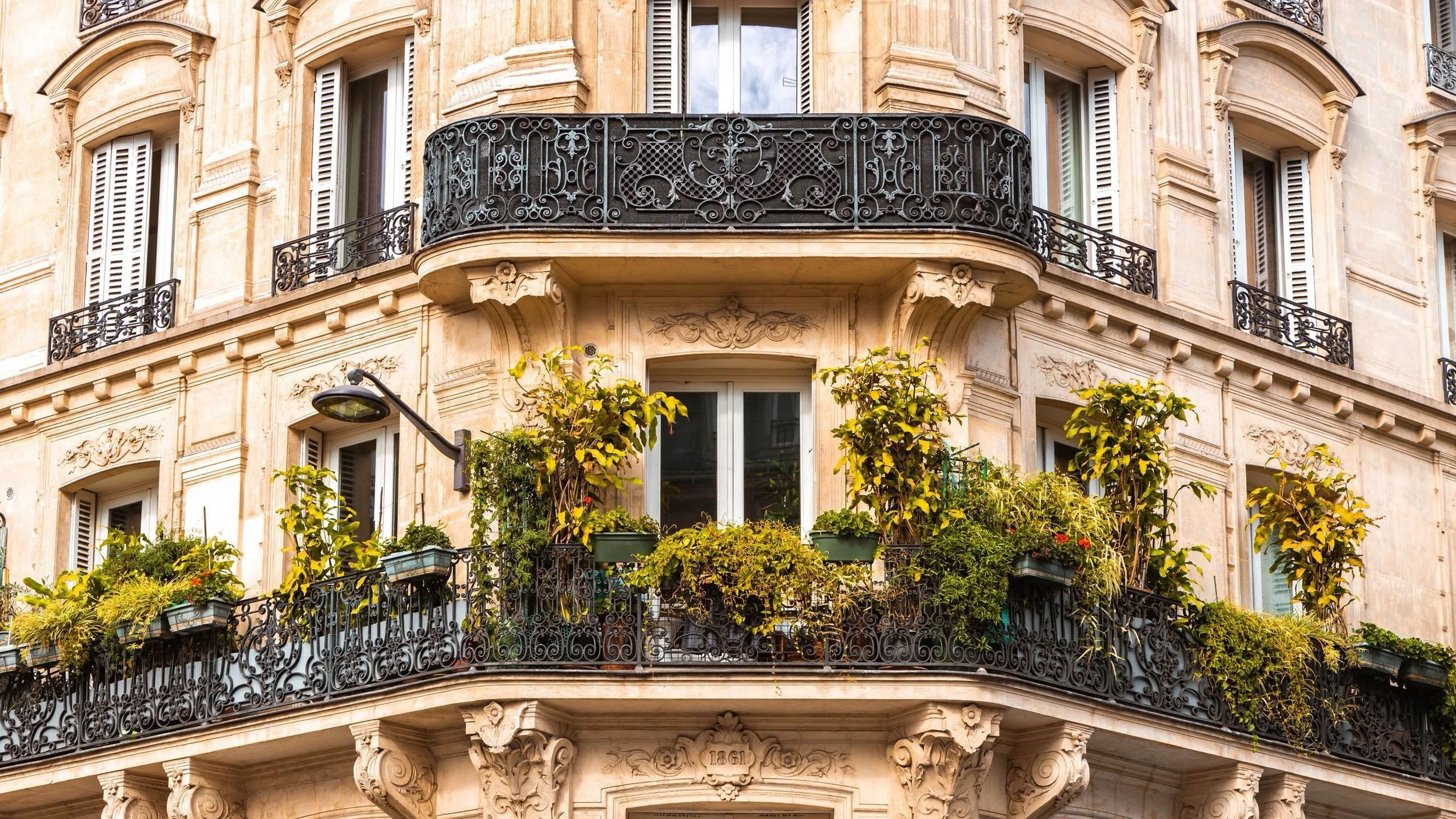 Façade d'un immeuble parisien avec balcons en fer forgé, fenêtres à volets, et façades en pierre gris clair, décorations ornementales et plantes vertes sur le balcon.