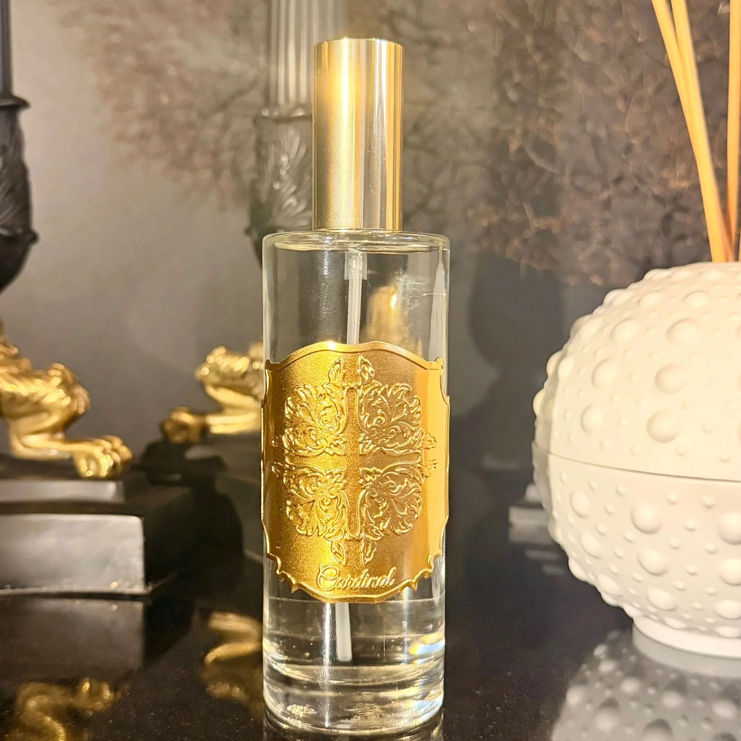 Un flacon de parfum transparent avec une étiquette dorée gravée, posé sur un comptoir noir. En arrière-plan, des bijoux dorés et une coque blanche sont visibles.