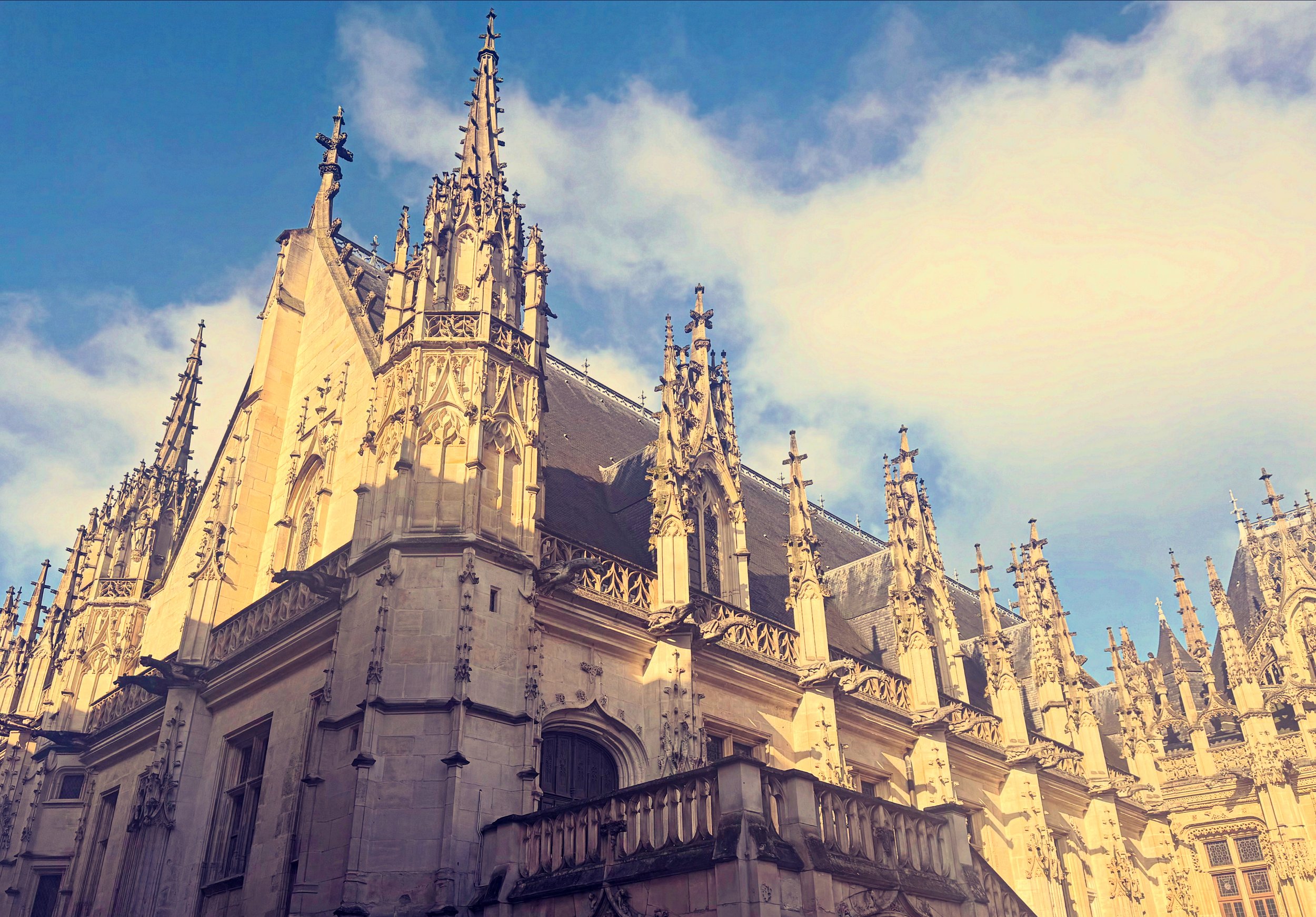 Une cathédrale gothique avec de hauts clochers ornés de pointes, en pierre claire, sous un ciel bleu avec quelques nuages.