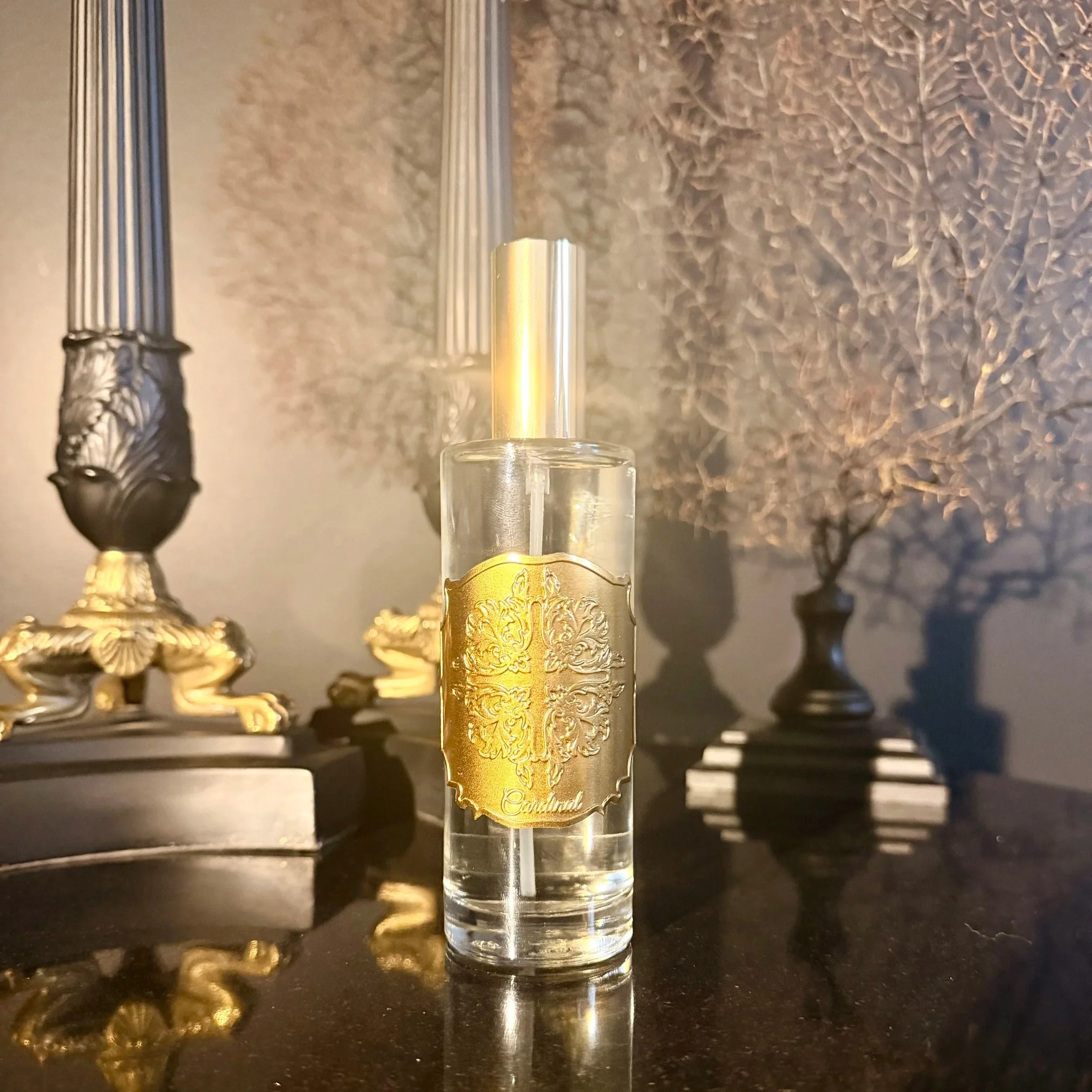 Une bouteille de parfum transparente avec une étiquette dorée décorative, positionnée au centre, devant un fond sombre avec des objets décoratifs en relief, notamment une gravure argentée et d'autres éléments en métal or.
