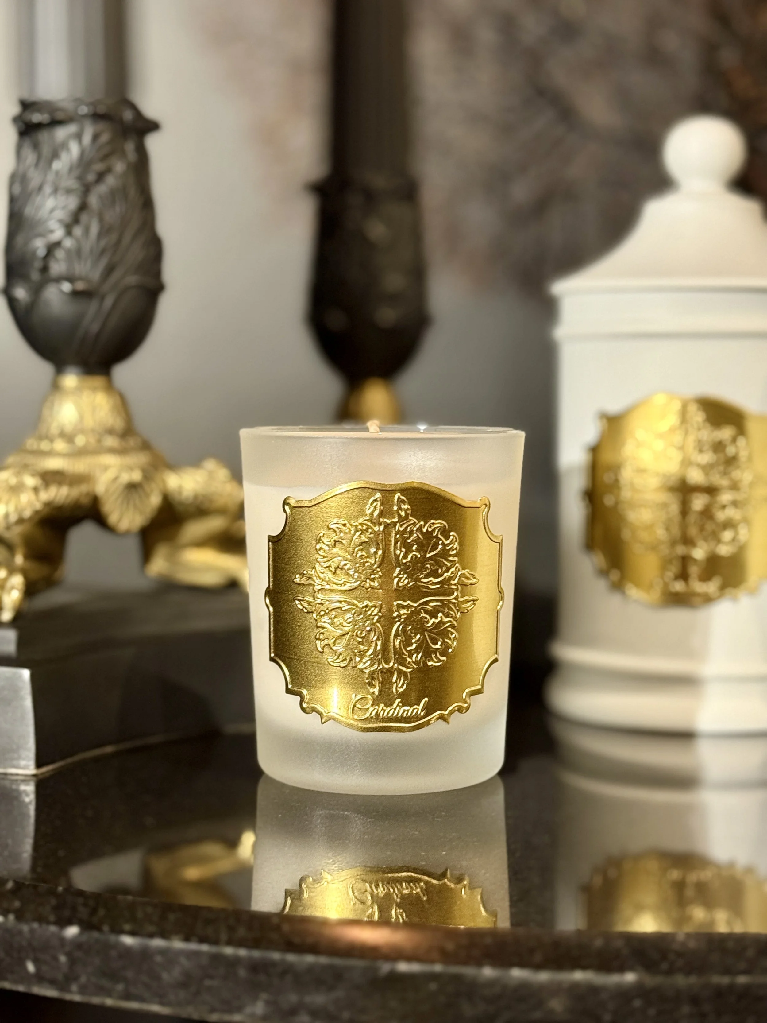 Bougie blanche ornée d'un label doré avec un motif d'un lion, placé sur une surface noire, avec d'autres objets dorés en arrière-plan.