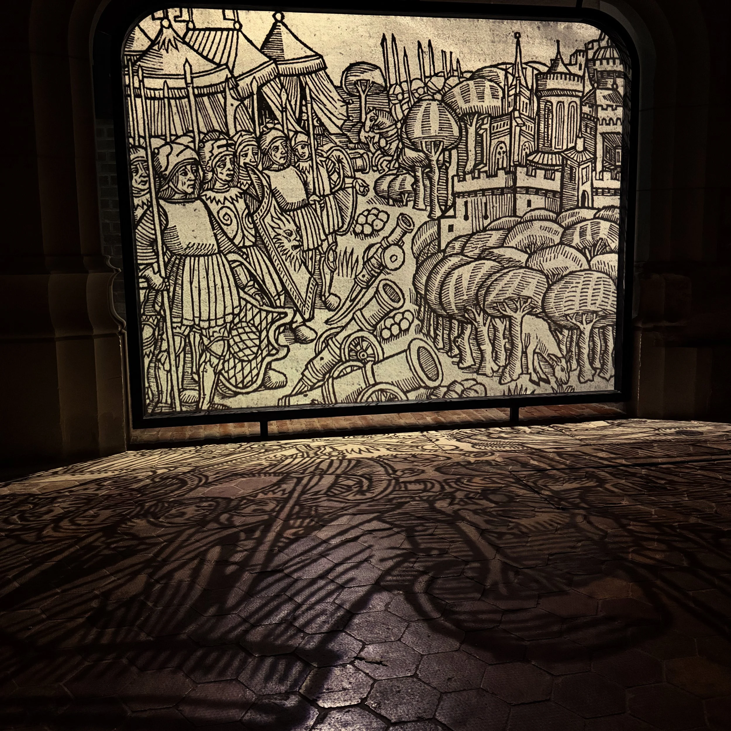 Une grande illustration noir et blanc représentant des personnages médiévaux, un château et des arbres, projetée sur un mur et un sol pavé.
