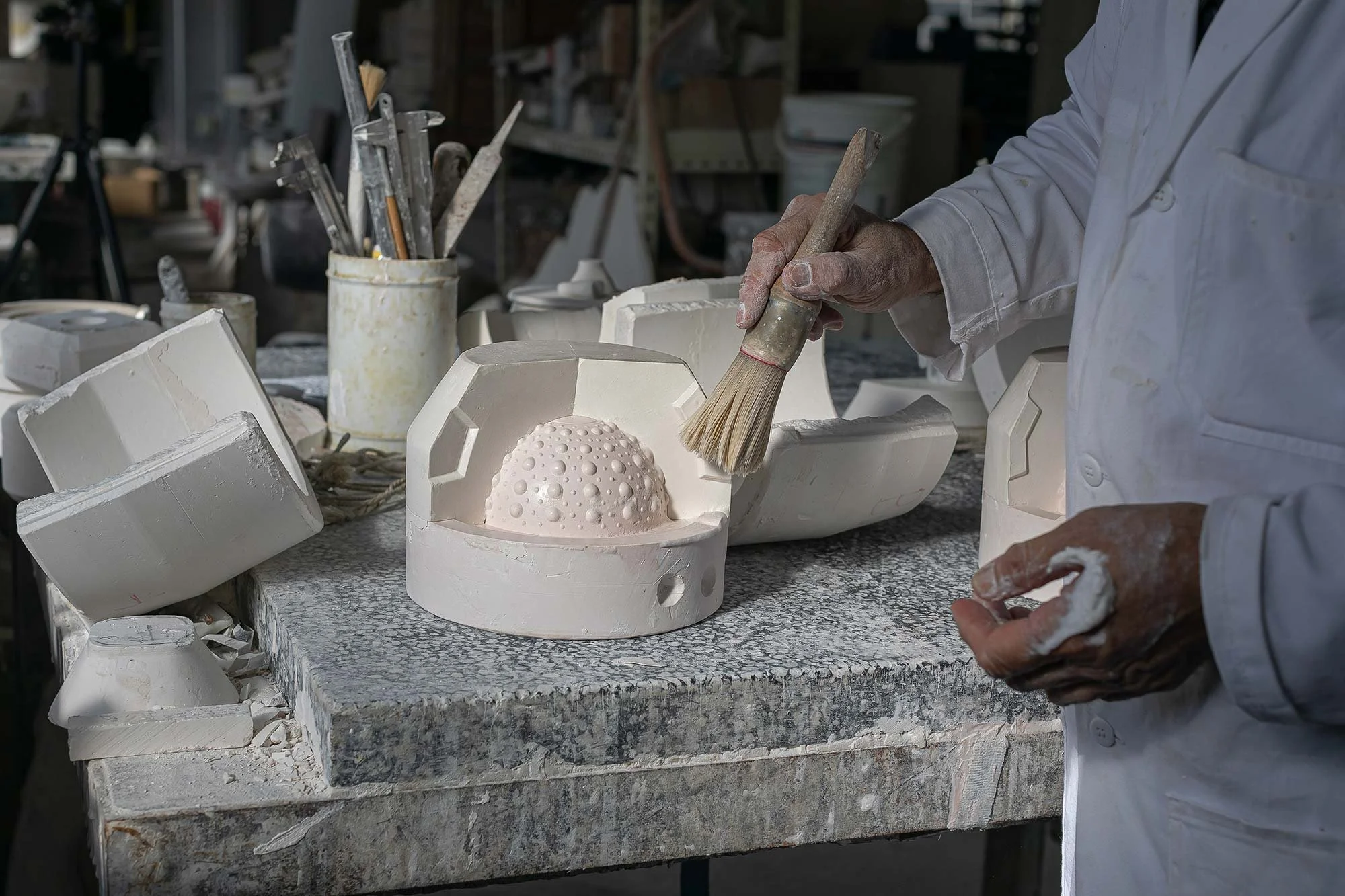 Un artisan en train de travailler le plâtre dans un atelier, entouré d'outils et de sculptures en cours de réalisation.