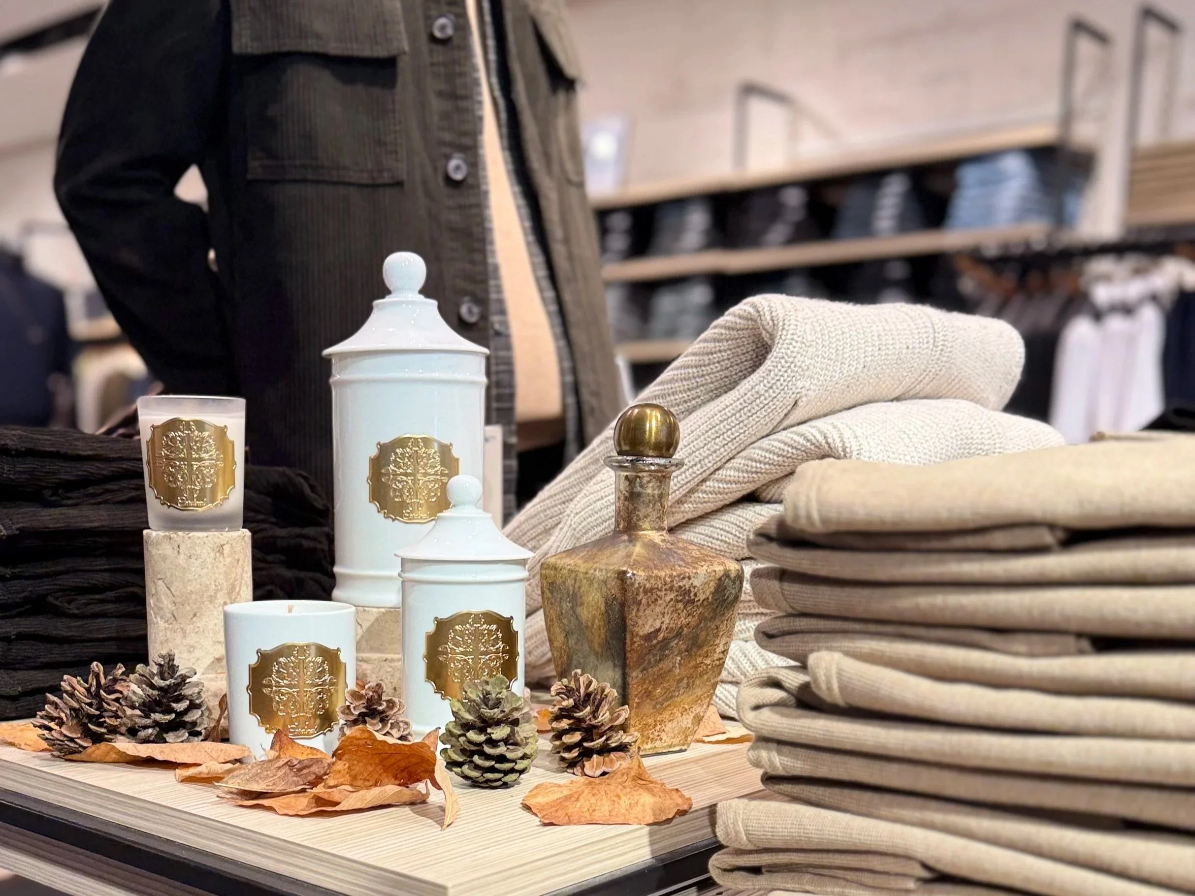 Présentoir de décoration avec bougies blanches ornementées de logos dorés, pommes de pin, branches d'automne, tissus en laine et objets en pierre, dans un magasin de mode ou décoration.