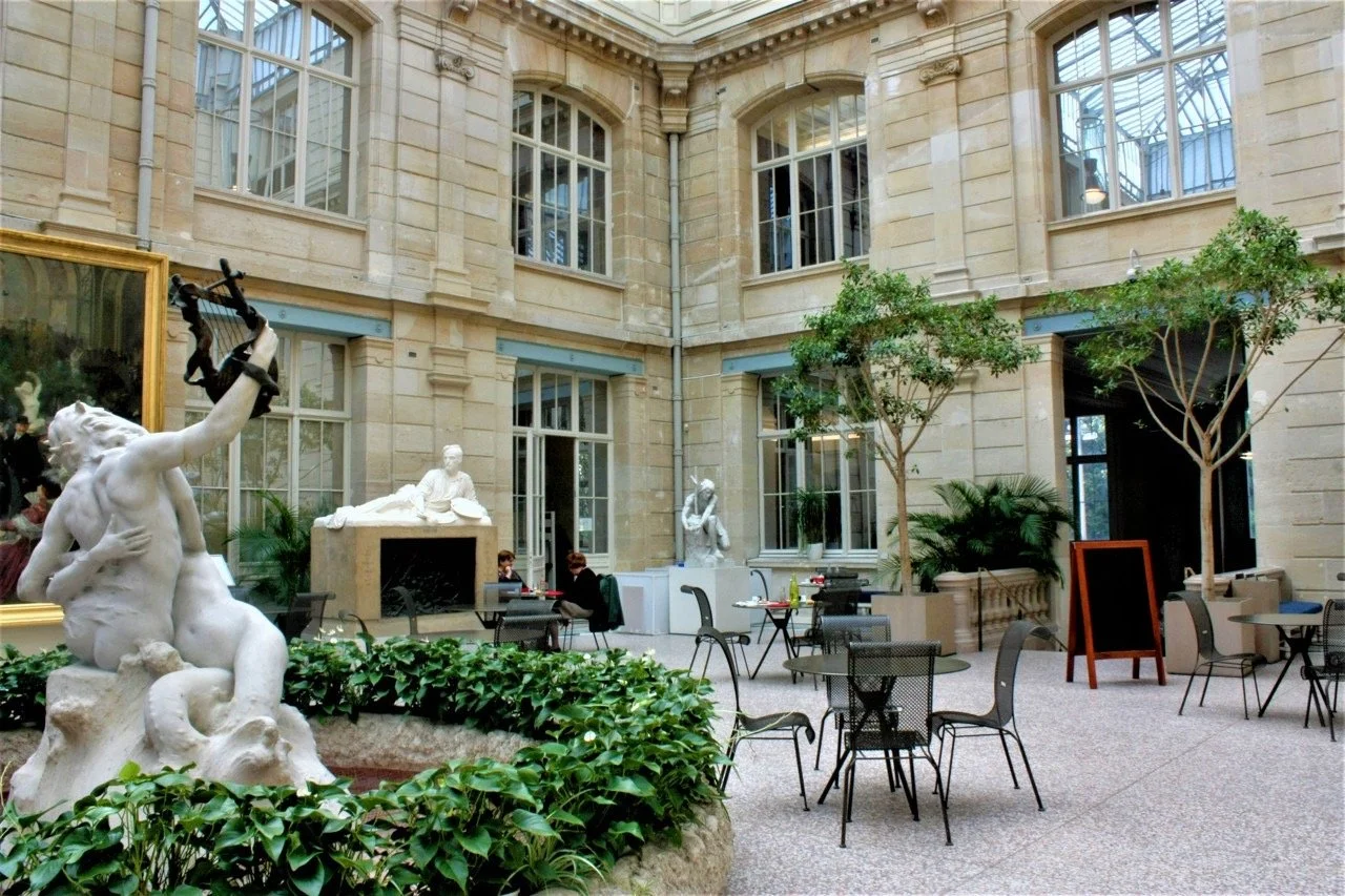 Intérieur d'une cour d'hôtel ou musée avec statues en marbre, plantes vertes, tables et chaises, grandes fenêtres avec cadre en métal, et un décor classique en pierre.
