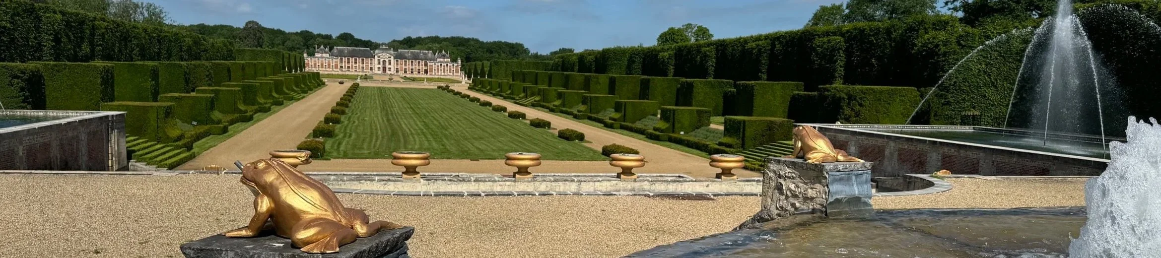 Un jardin à la française avec des fontaines ornées de grenouilles en or et un château en arrière-plan.