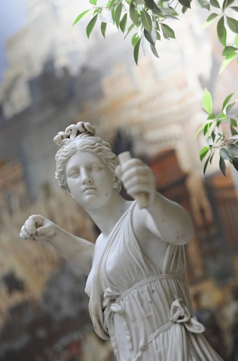 Sculpture en marbre représentant une femme avec une couronne de tresses, tenant une épée, dans un environnement intérieur avec des plantes et une vieille muraille en arrière-plan.