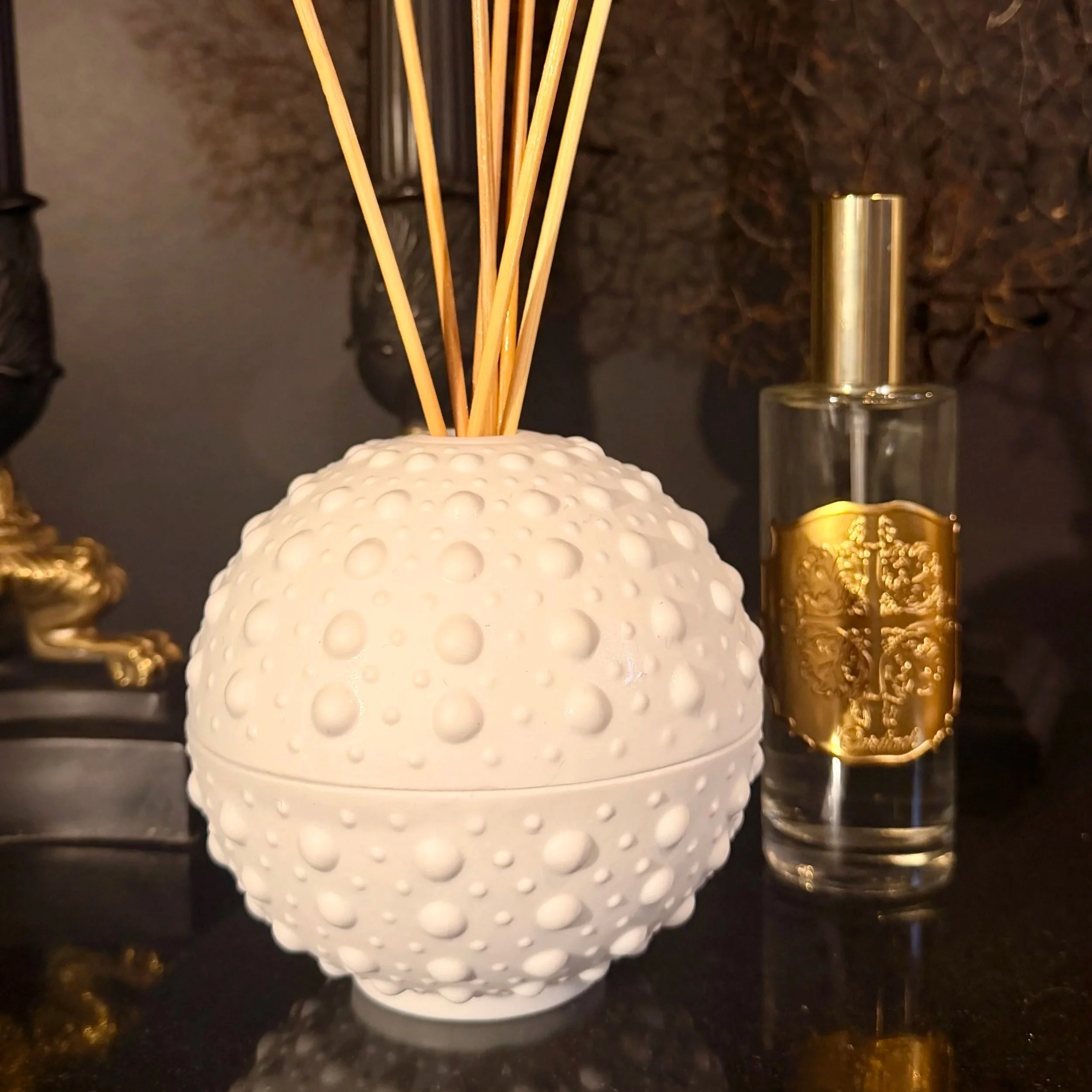 Un diffuseur d'huiles essentielles blanc avec une surface textured et plusieurs bâtons en bois à l'intérieur, placé sur une surface noire avec un décor doré et noir en arrière-plan.
