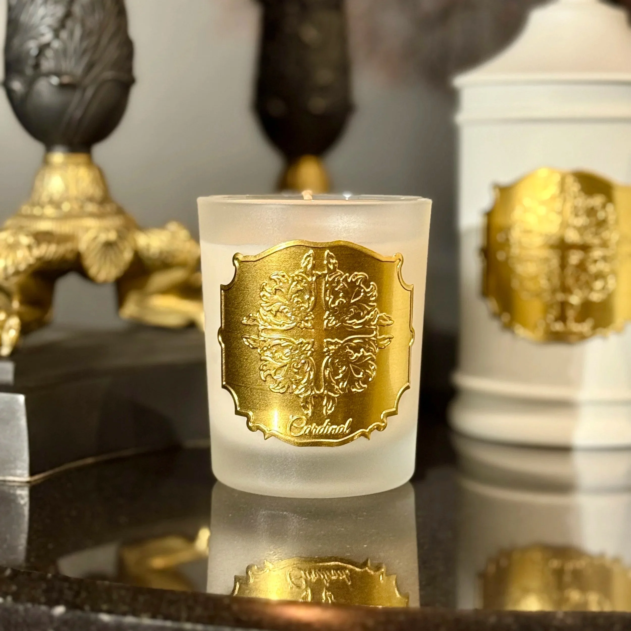 Une bougie blanche avec un label doré portant un motif de vigne et le nom 'Cardinal', entourée de décorations dorées et de vases noirs en arrière-plan.