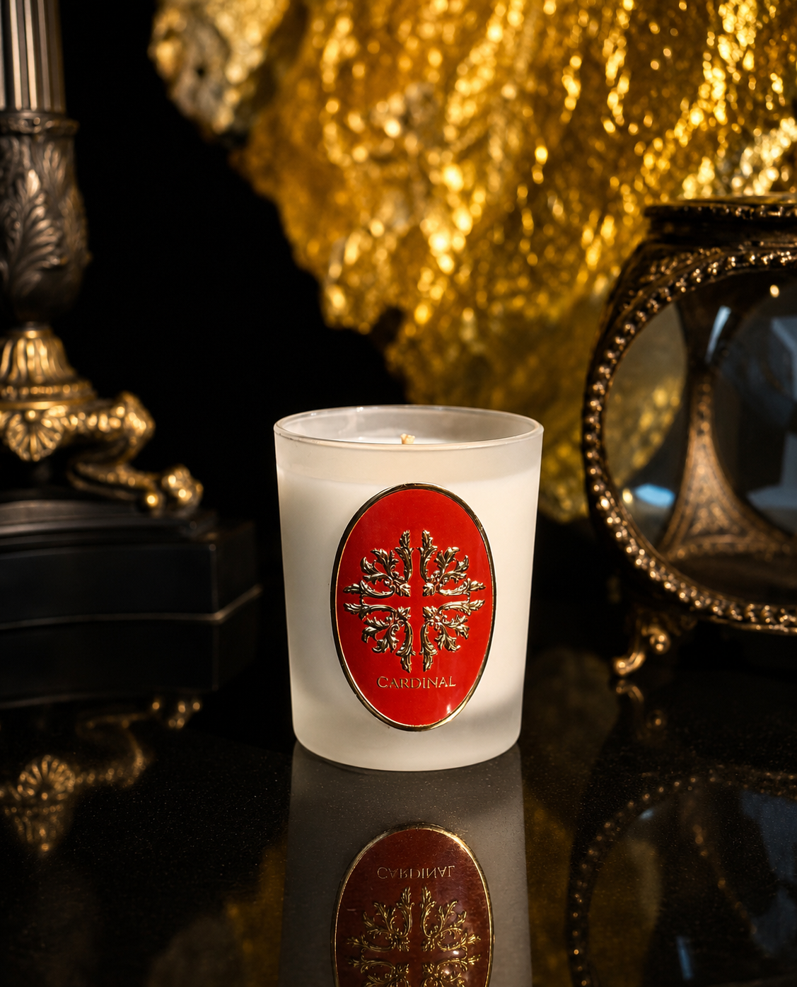 Une bougie blanche avec un label doré portant un motif de vigne et le nom 'Cardinal', entourée de décorations dorées et de vases noirs en arrière-plan.