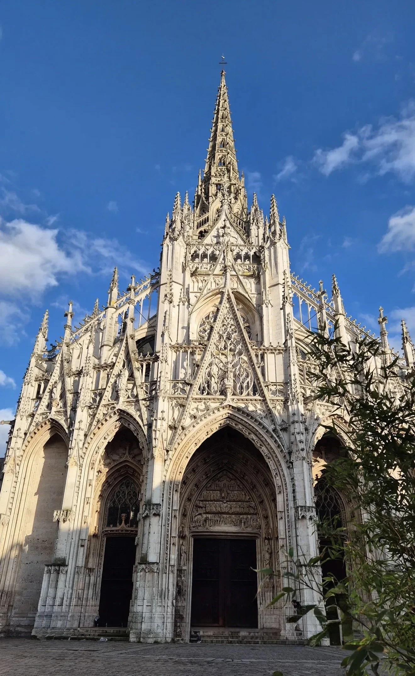 Façade gothique de la cathédrale avec une entrée ornée et une flèche élancée vers le ciel, sous un ciel bleu avec quelques nuages.