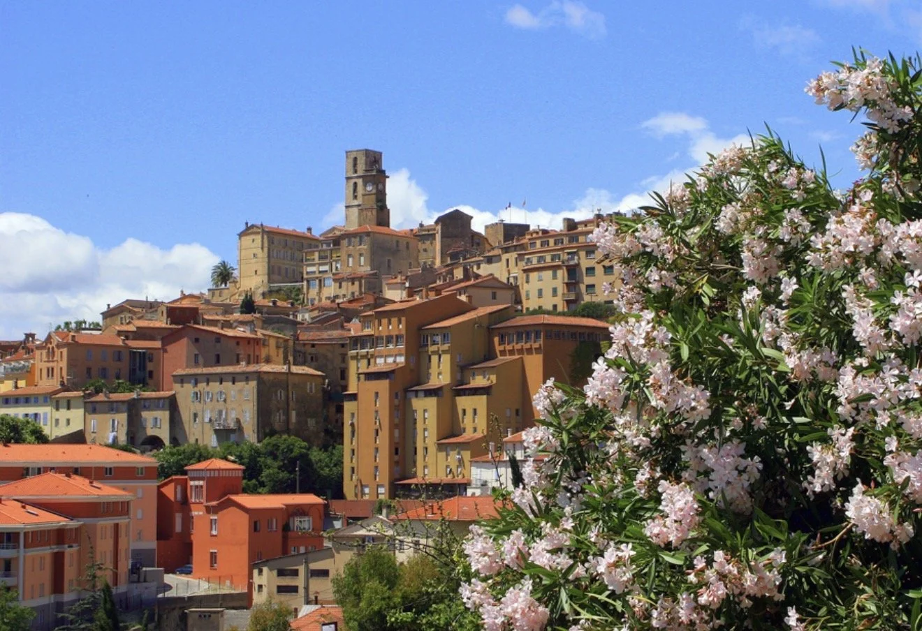 Ville perchée avec des bâtiments colorés et un clocher, entourée de fleurs roses et de végétation, sous un ciel bleu avec quelques nuages.