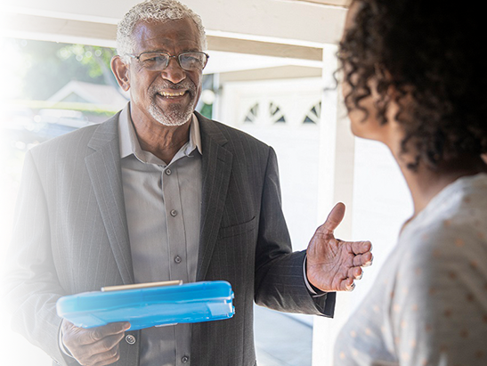 An elderly man with grey hair and glasses, wearing a suit, is smiling and talking to a woman with curly hair, who is partially visible. They are outdoors or on a porch.