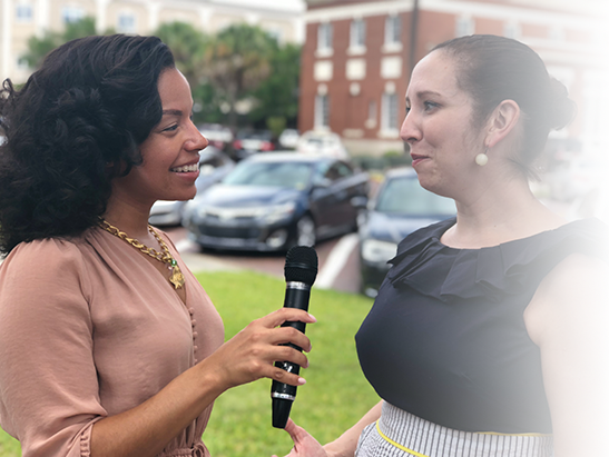 Two women interviewing each other outdoors; one holds a microphone, and they are smiling at each other.