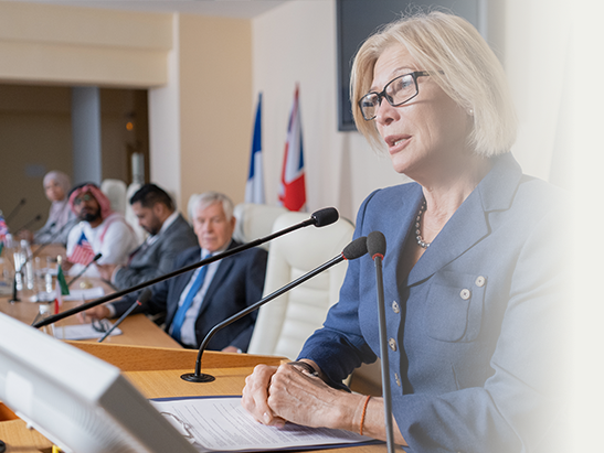A woman in a blue blazer speaking at a panel with multiple microphones, in a conference room with other diverse people and flags in the background.