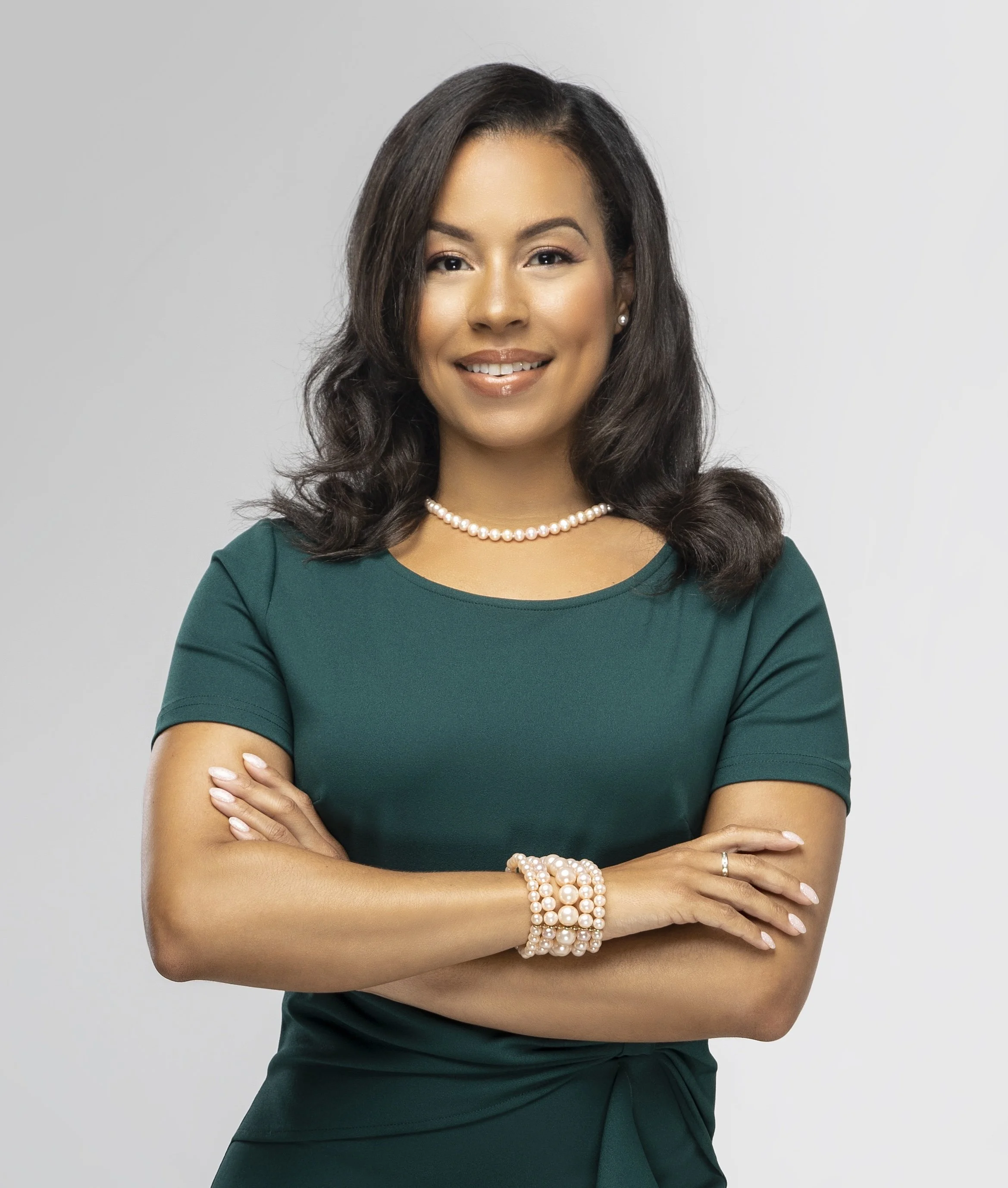 A woman with shoulder-length dark hair, wearing a green dress, pearl necklace, bracelet, and earrings, standing with arms crossed and smiling against a plain gray background.