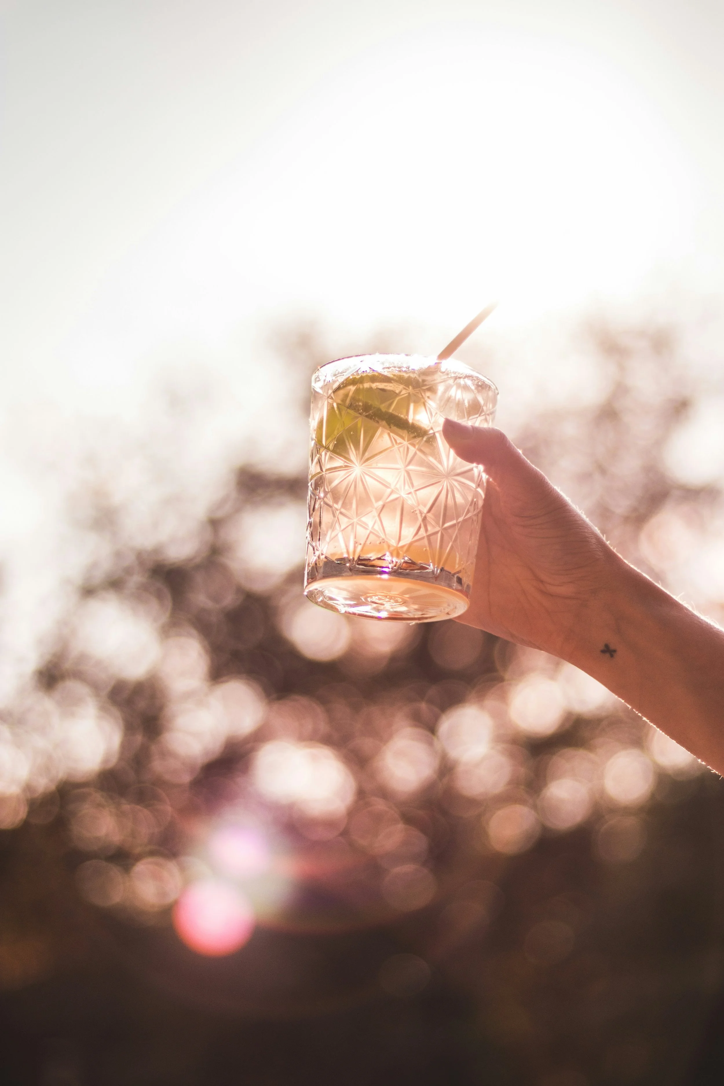 Hand holding a cocktail in a patterned glass with lime and straw against a soft-focus, sunlit background.