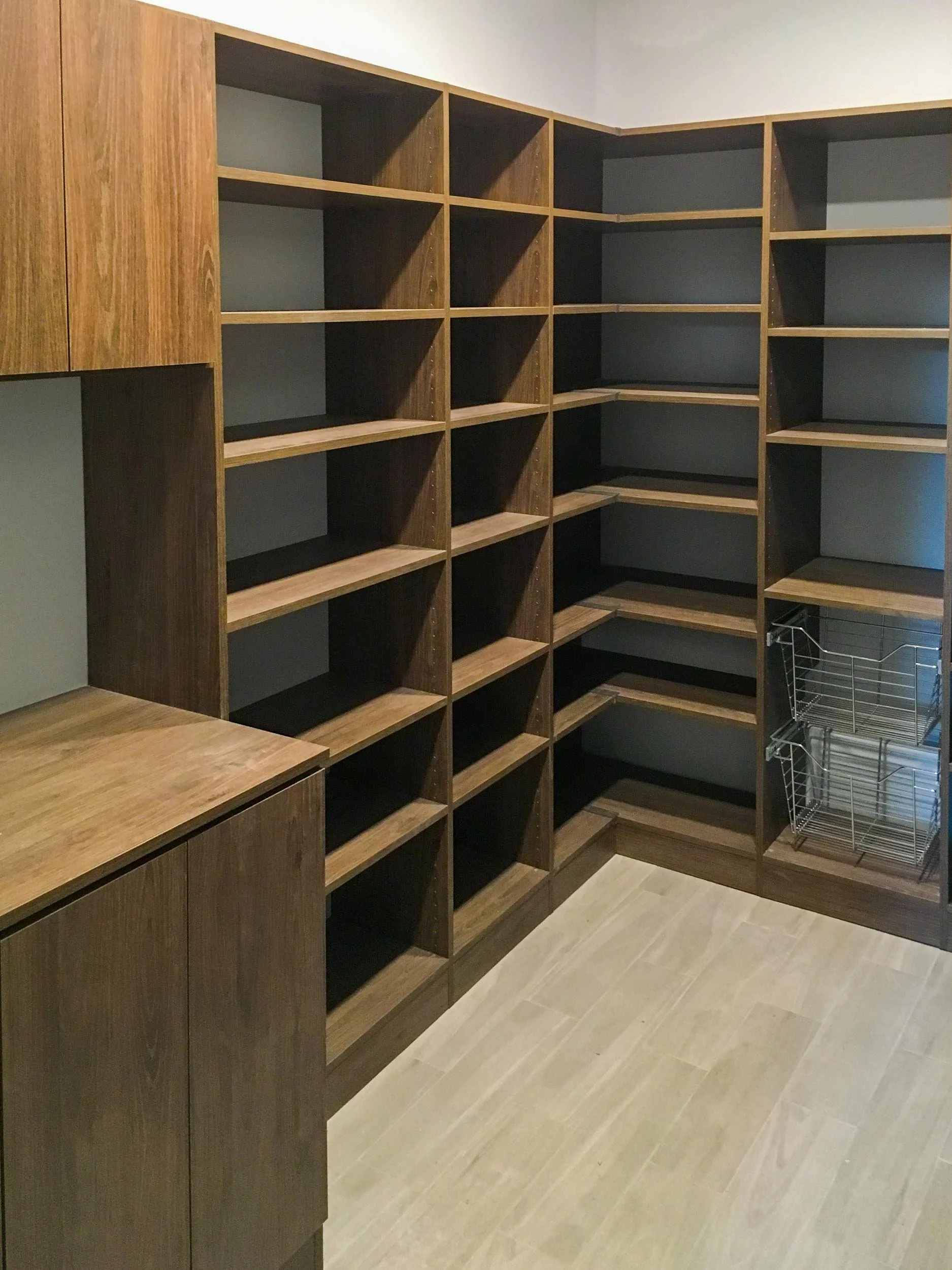 Empty walk-in closet with brown wooden shelves and cabinets, and a wire storage basket on a light gray tiled floor.