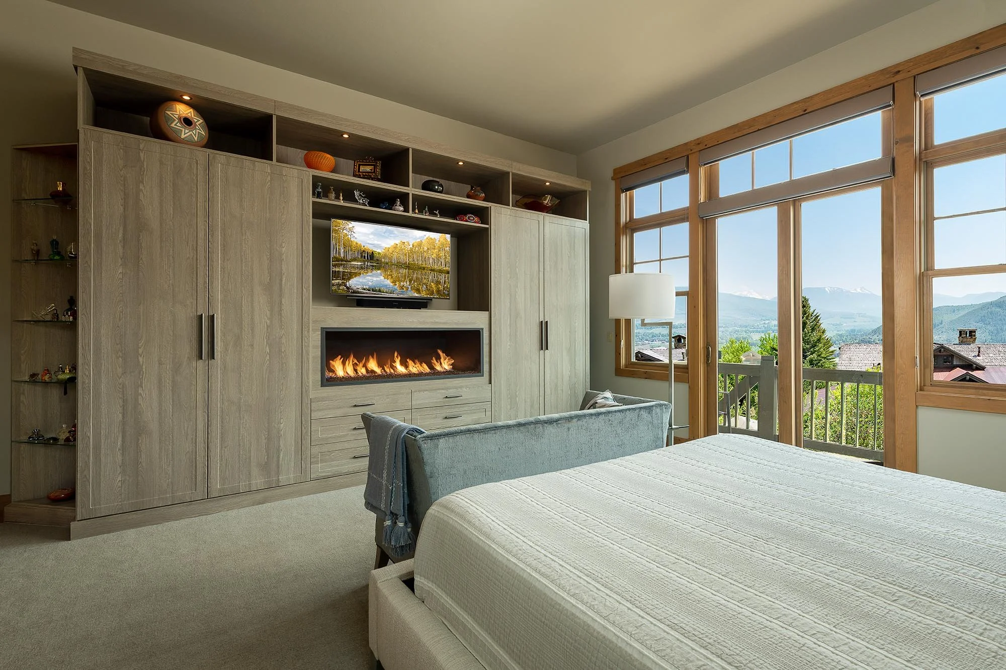 A bedroom with large windows showing a scenic mountain view, a white bed, a TV above a fireplace, and light wood furniture.