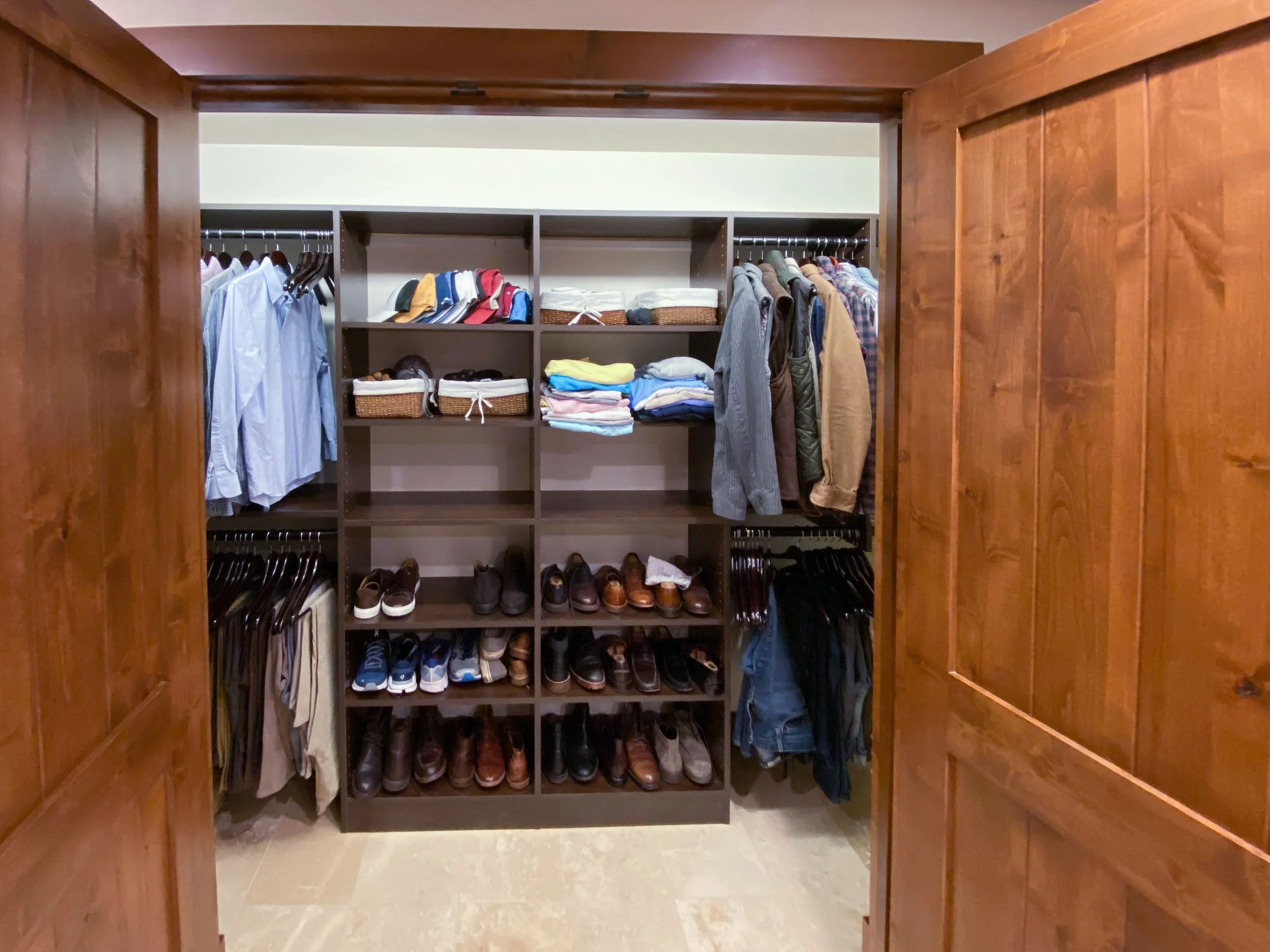 View of a well-organized closet with a dark brown shelving unit containing folded clothes, shoes, hats, and jackets. Two wooden closet doors are partially open, revealing the interior.
