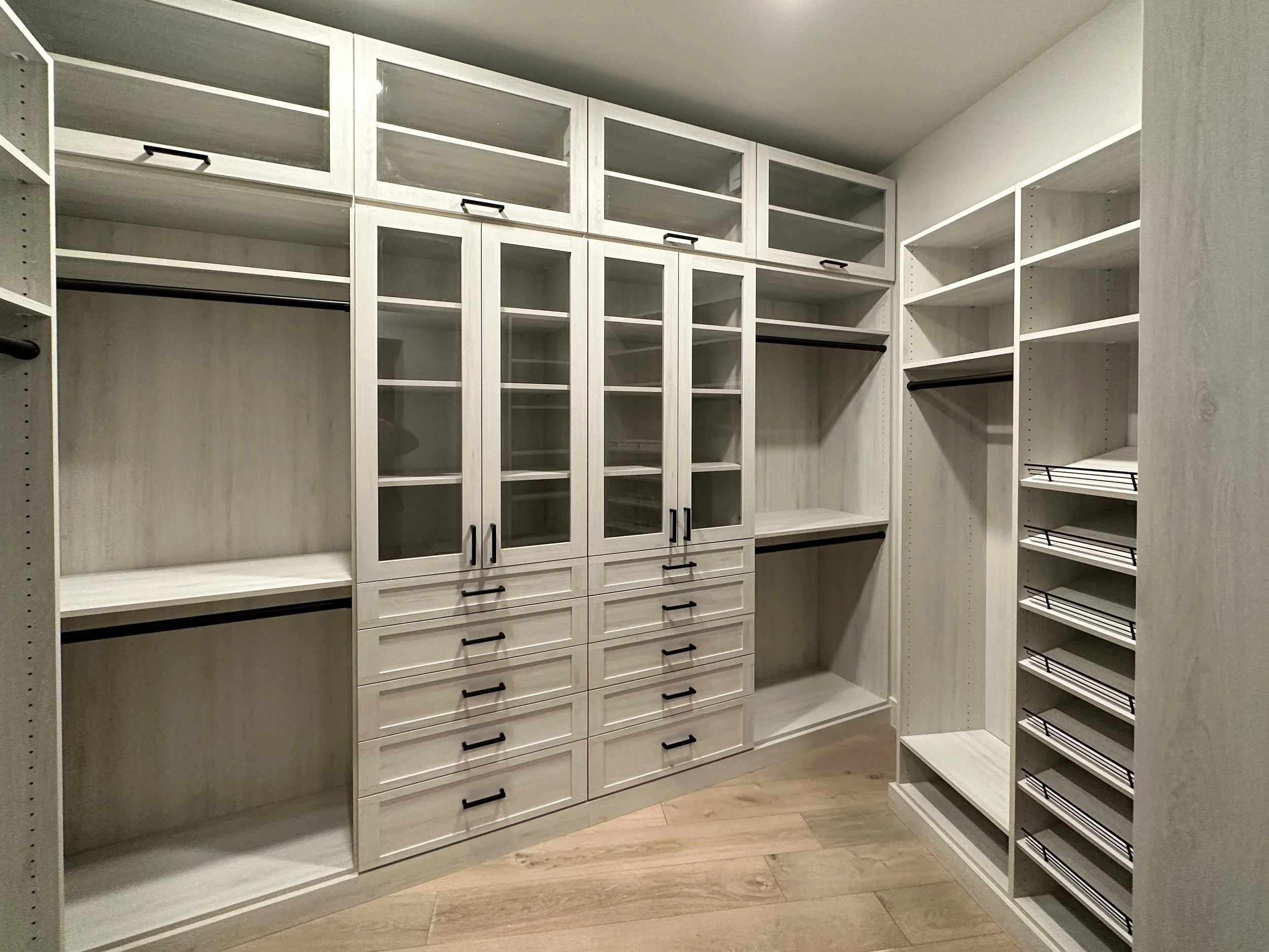Empty white closet with glass-paneled cabinets, drawers, and multiple shelving units, with some black hanging rods and wire baskets, on a wooden floor.