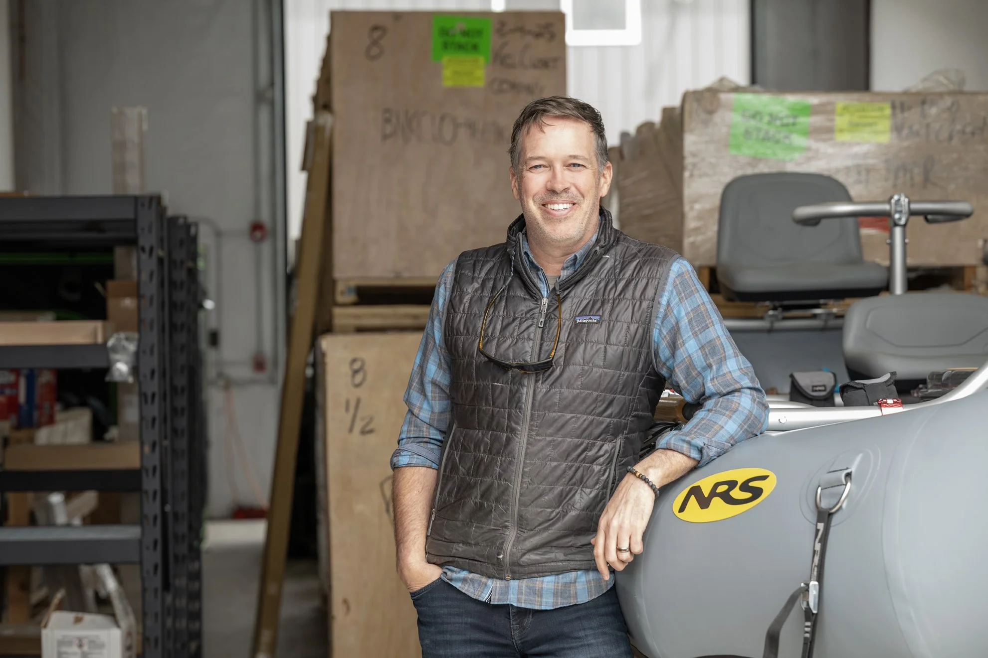 Owner Brian Bloess,  smiling, wearing a gray vest over a plaid shirt, standing in a warehouse with boxes and equipment around him.