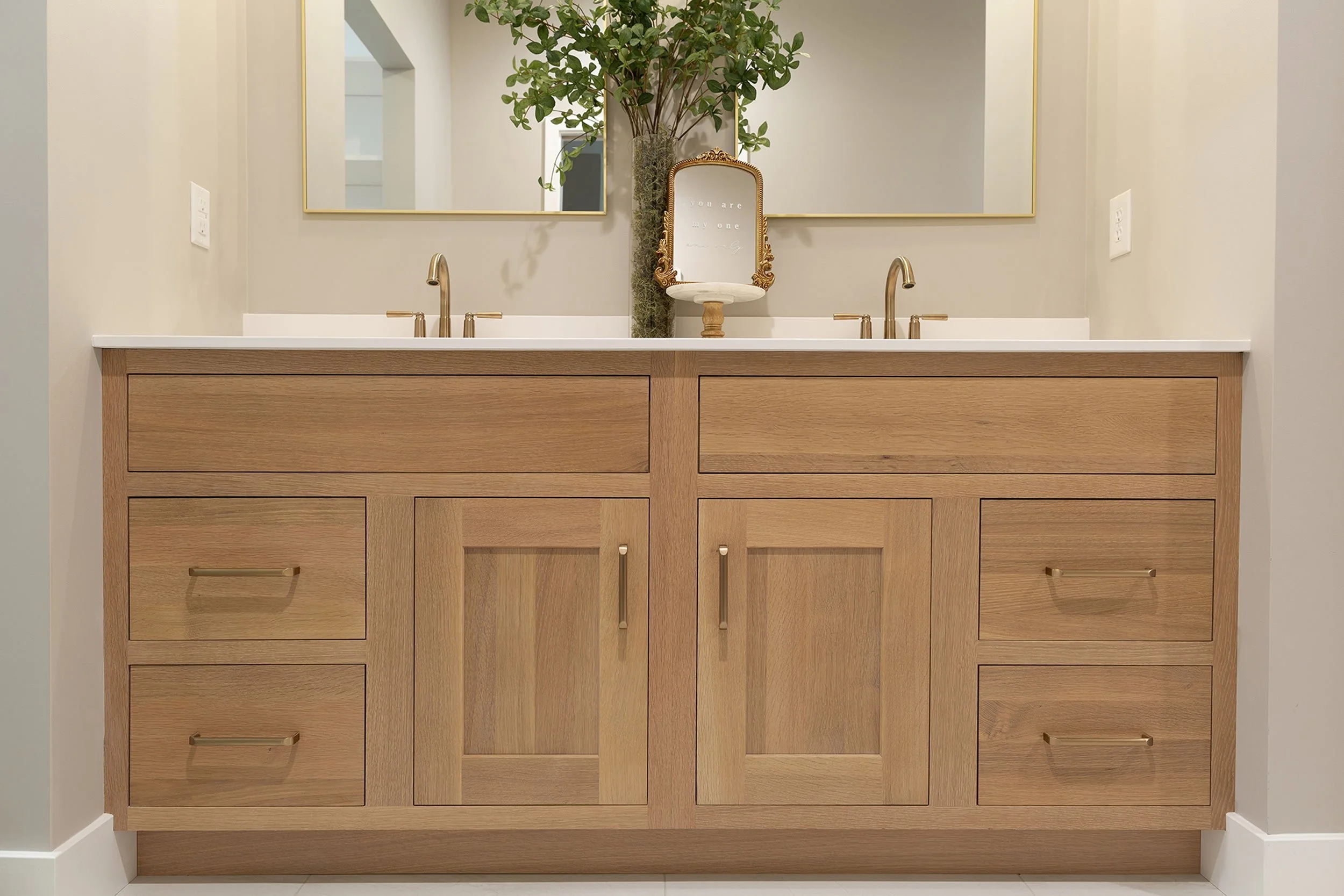 A double bathroom vanity with a large mirror above, two gold faucets, a decorative framed mirror, and a tall vase with greenery inside. There is a small framed quote on the chair that says, 'You are my one.'