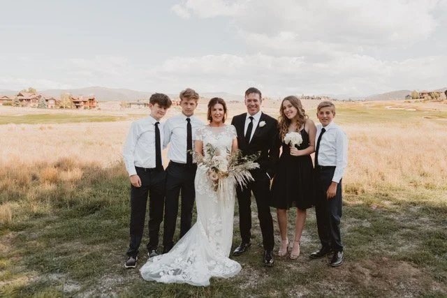 Owner Brian Bloess and six family members, including a bride and groom, standing outdoors in a grassy field with houses and mountains in the background.