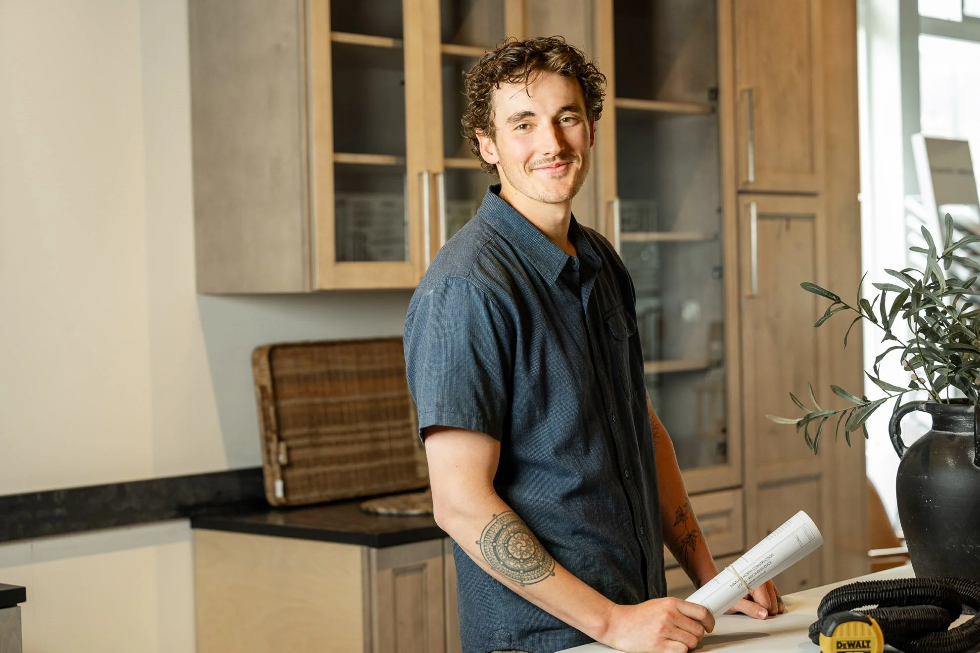A young man with curly hair and tattoos on his arm standing in a modern kitchen, holding a rolled-up blueprint or plan, smiling at the camera.