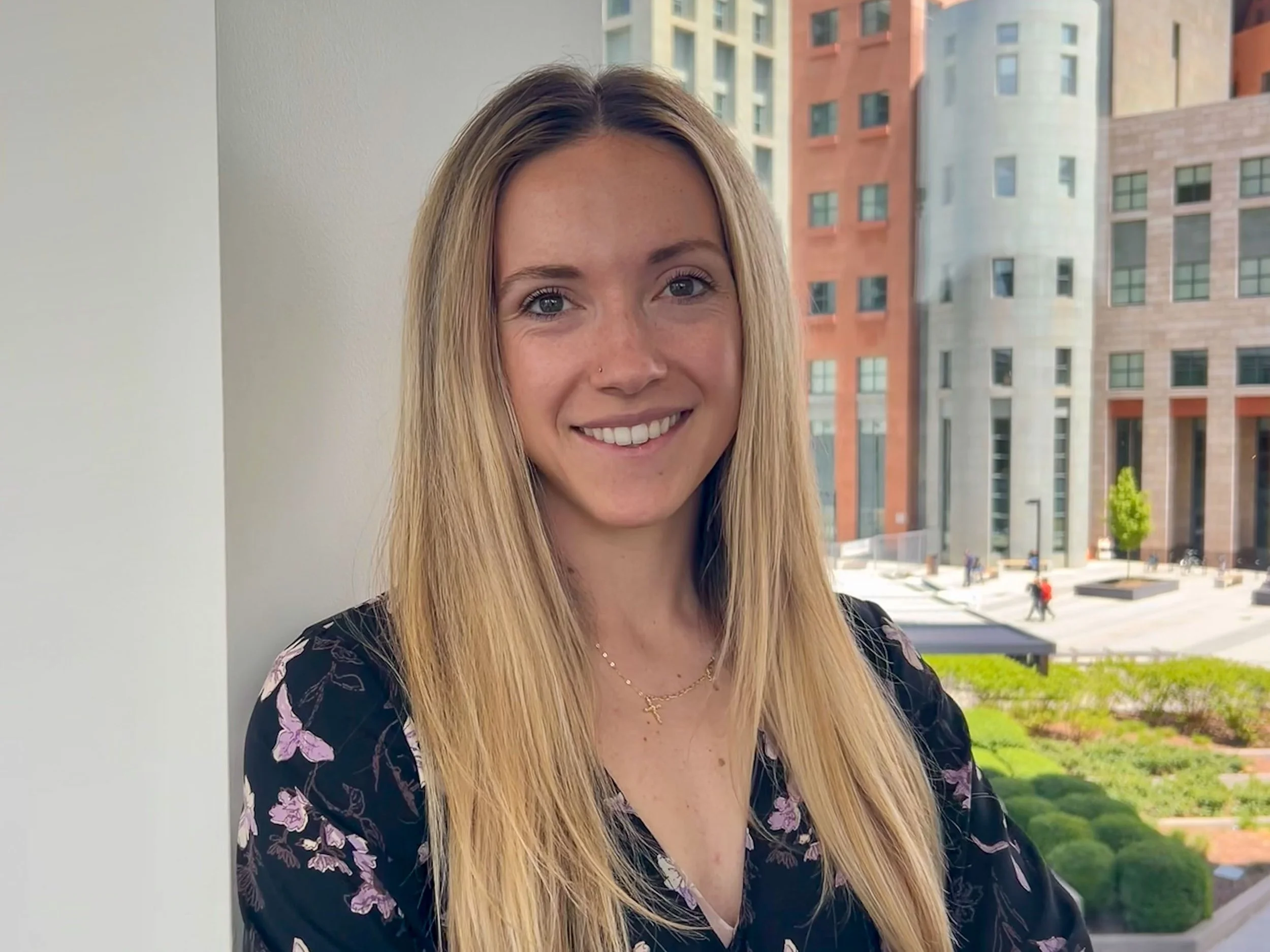 A woman with long blonde hair, wearing a floral top and a necklace, smiling while standing outdoors with a cityscape of modern buildings in the background.