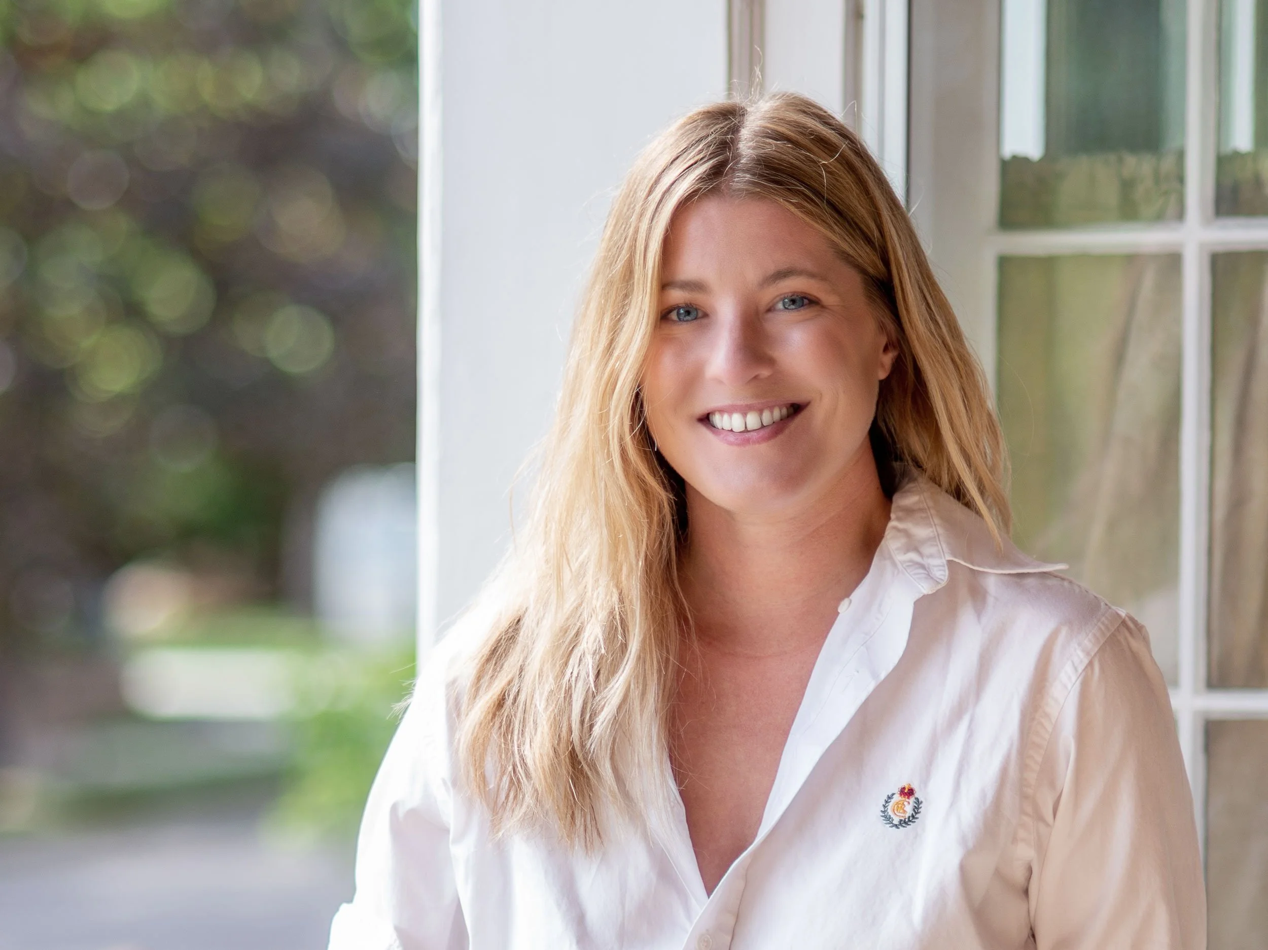 A woman with blonde hair and blue eyes smiling, wearing a white shirt with a small embroidered logo on the chest, sitting by a window.