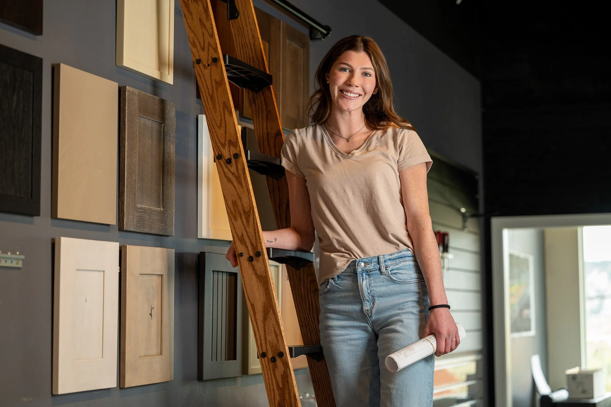 A woman with shoulder-length brown hair wearing a beige t-shirt and blue jeans smiles while standing next to a wooden ladder in a showroom with various cabinet samples on the wall.