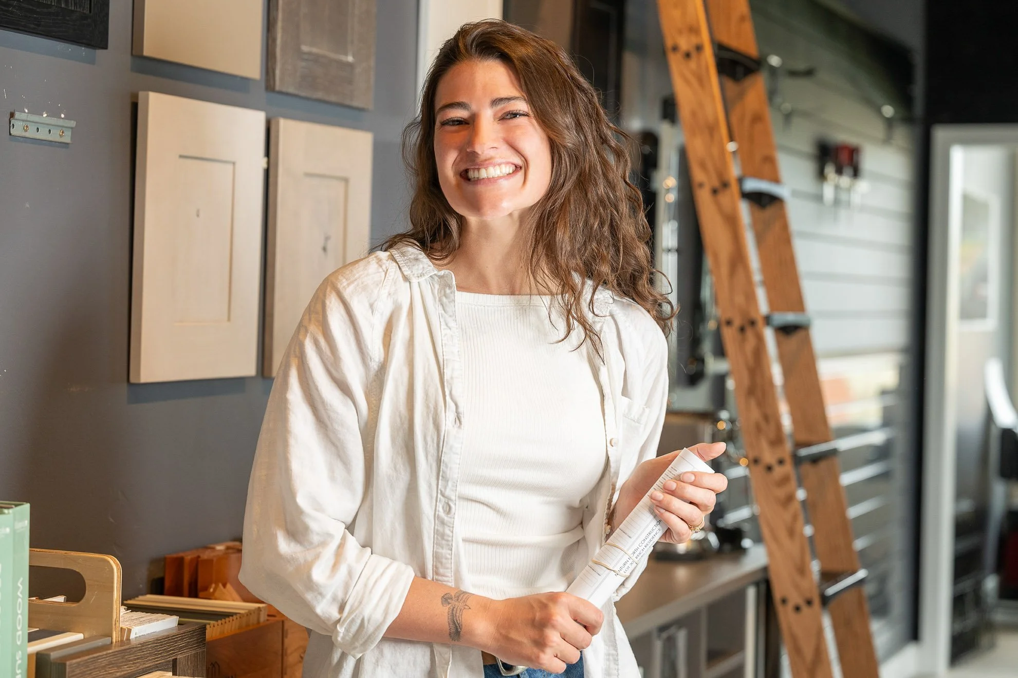 A smiling woman with long brown hair, wearing a white shirt, holding a rolled-up paper, standing inside a store or showroom with artwork and wooden ladders on the walls.