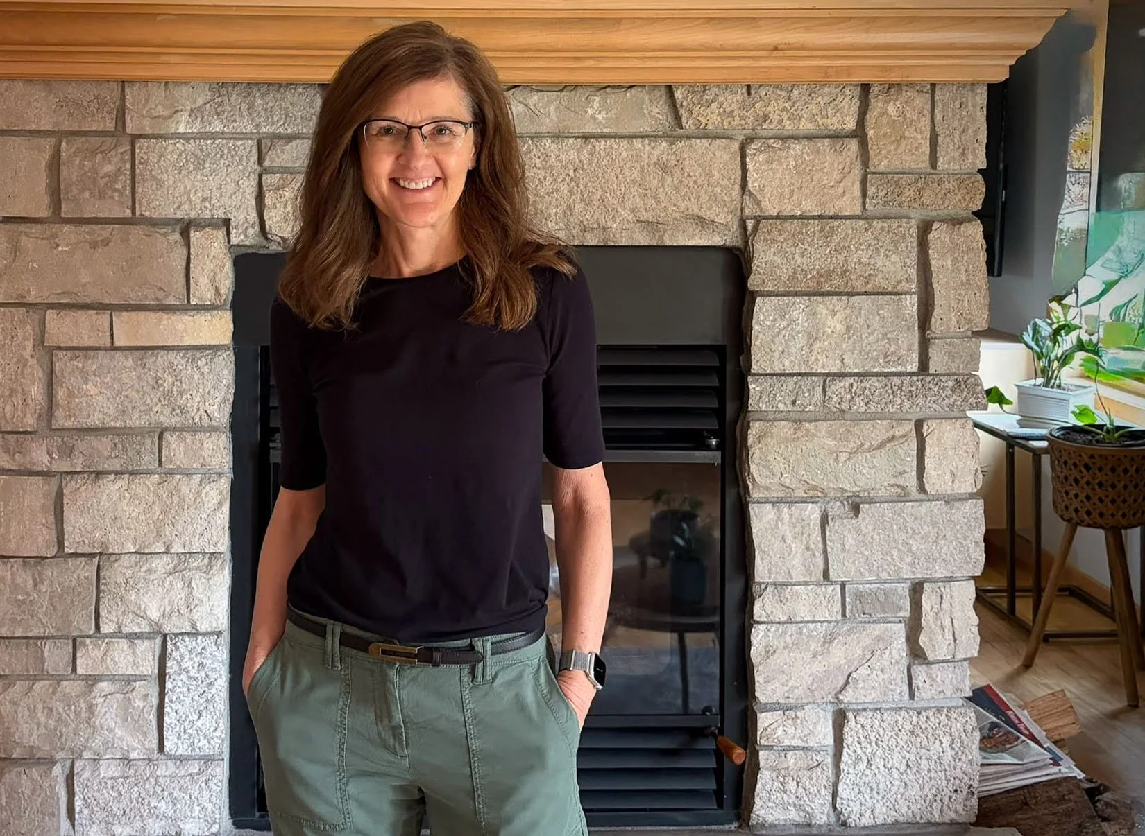 Mary Liegel with brown hair, glasses, and a black shirt standing in front of a stone fireplace with some green plants and a window on the right side.