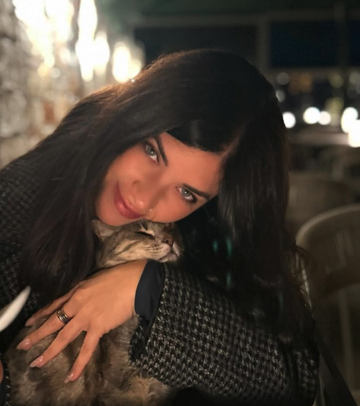 Woman with long dark hair cuddling a tabby cat indoors.