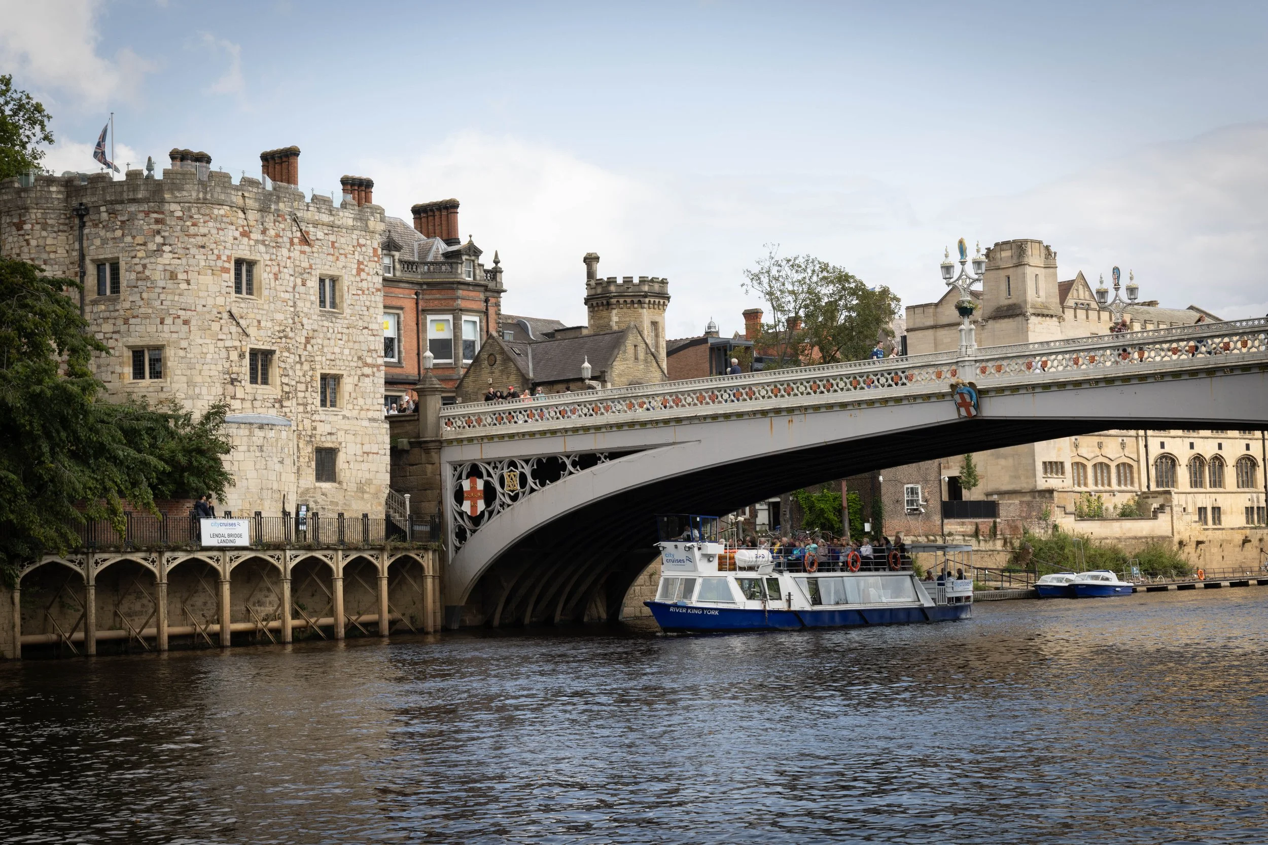 View of the River Thames with a boat named River King York, historic buildings and a bridge in London, England, with tourists walking on the bridge and a Union Jack flag flying.