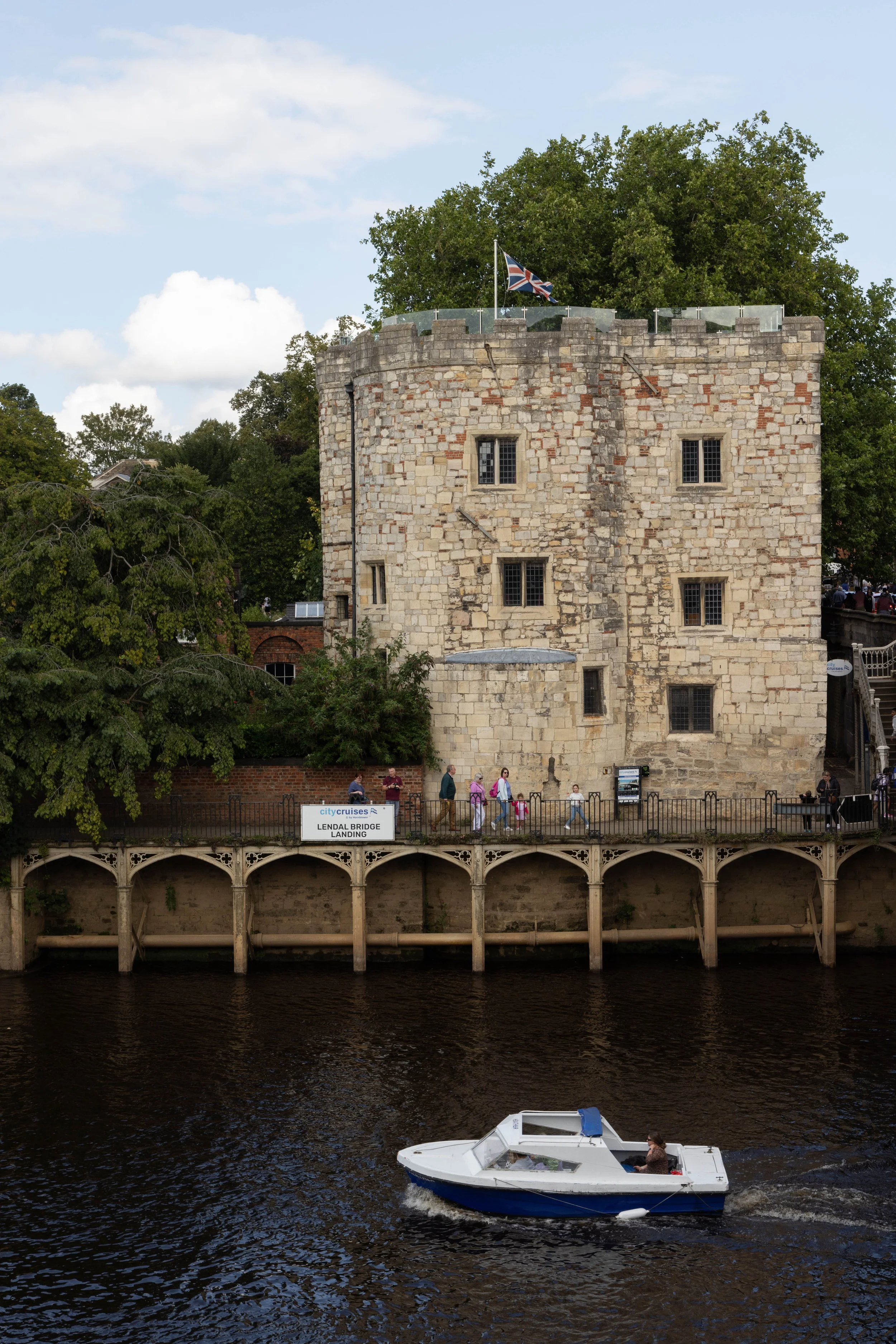 A historic stone tower with windows and a Union Jack flag on top, located beside a river. People are walking along the riverbank, and a small boat with a woman on it is on the water below. There is a sign that reads 'Lendal Bridge Landing'.