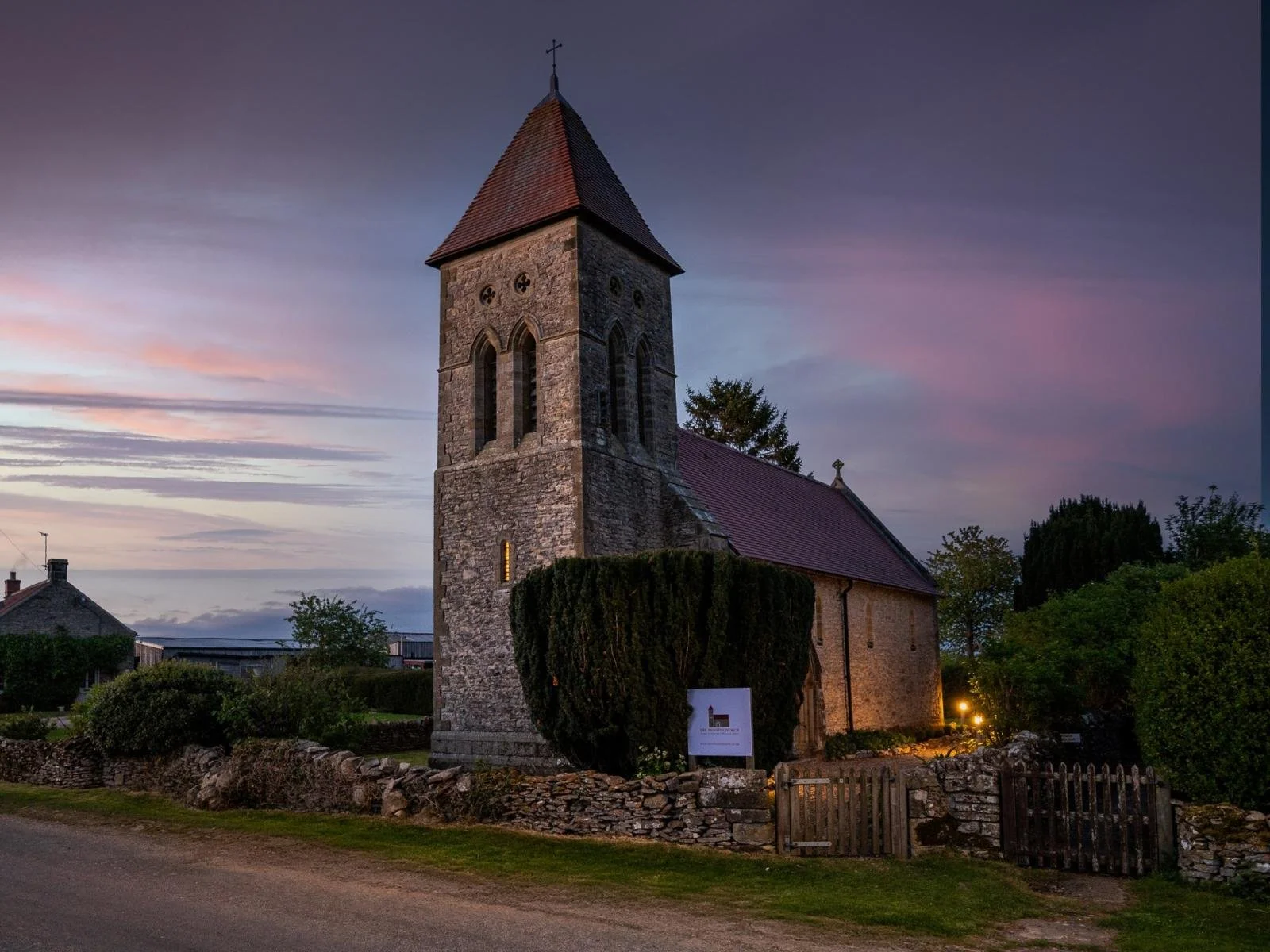 A stone church with a tall steeple during sunset, surrounded by trees and a low stone fence.