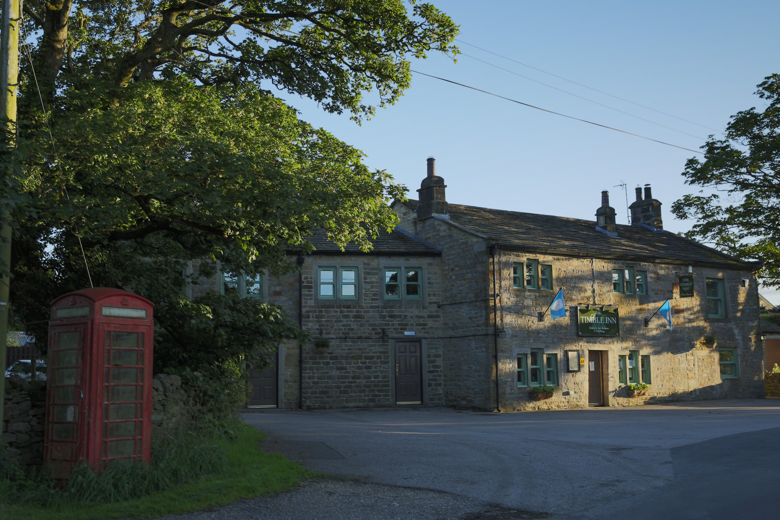 A stone building with a sign that reads 'Timble Inn' on the front, surrounded by green trees, with a traditional red British telephone box near the road in front of it during what appears to be late afternoon or early evening.