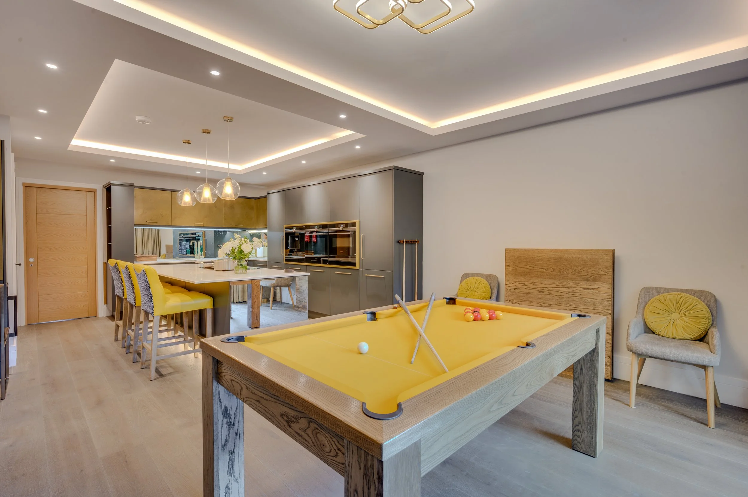 Modern living room with yellow pool table, pool cues, and ball rack, featuring a kitchen in the background with gray cabinets, a white marble island, and decorative lighting on the ceiling.