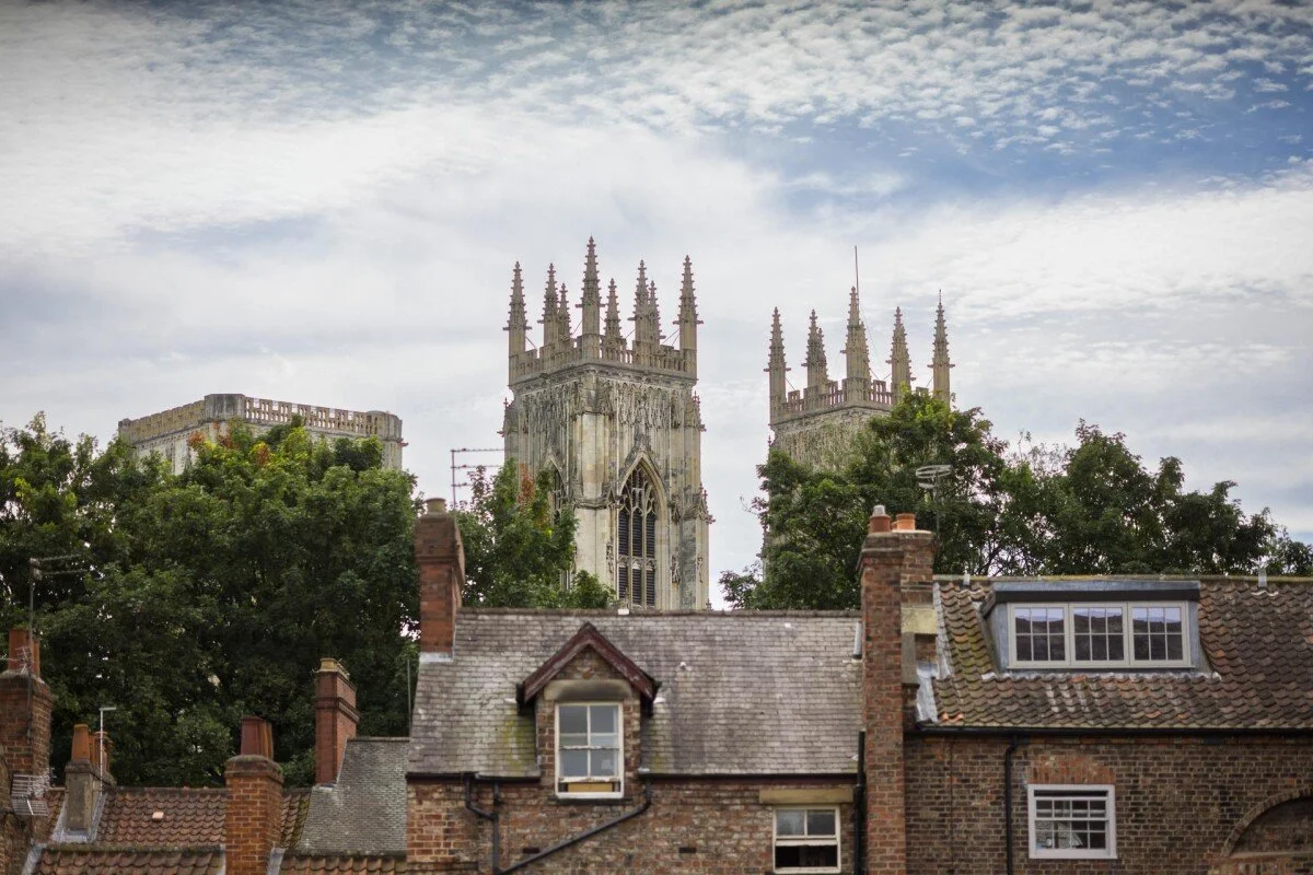 A historic church with gothic architecture and tall spires, partially obscured by trees and traditional brick houses in the foreground under a cloudy sky.