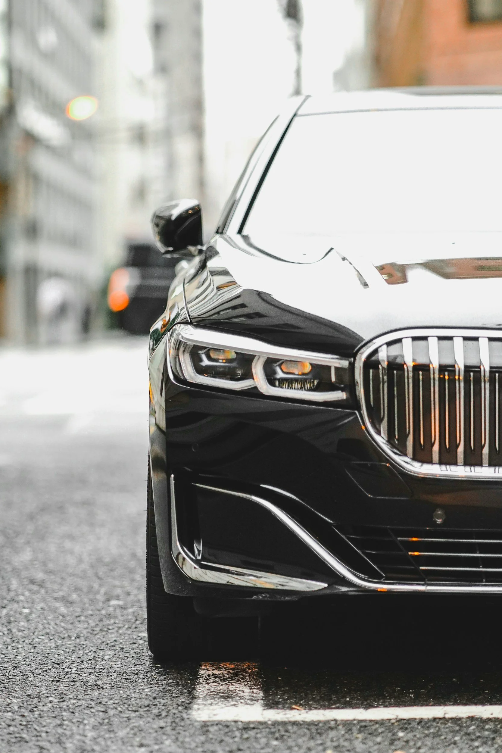Close-up of the front right side of a black luxury car, showing the headlight, grille, and side mirror, parked on a wet urban street.