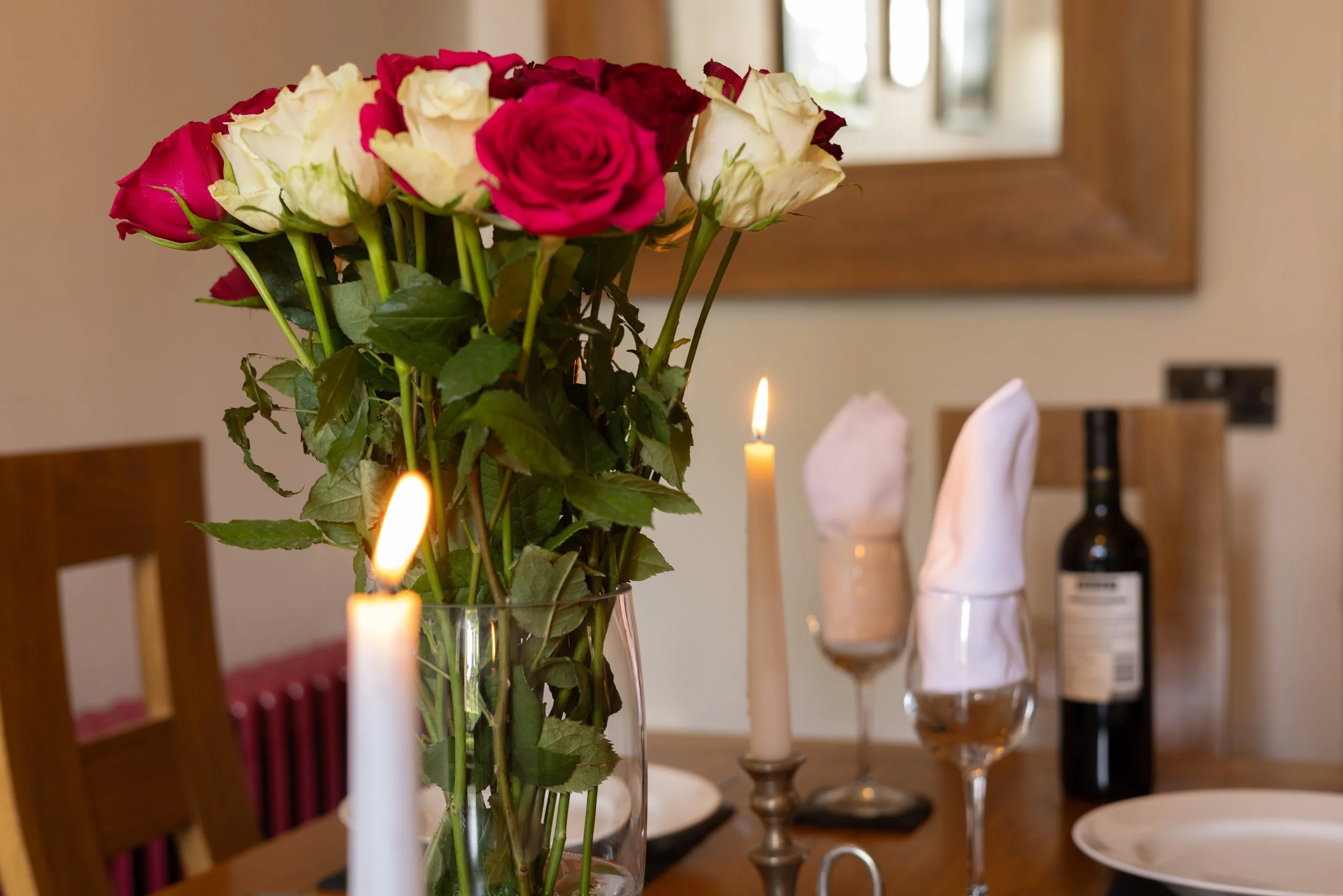 A vase of mixed pink and white roses on a dining table. The table is decorated with white candles and set with plates and glasses. In the background, there is a wooden-framed mirror and a bottle of wine.