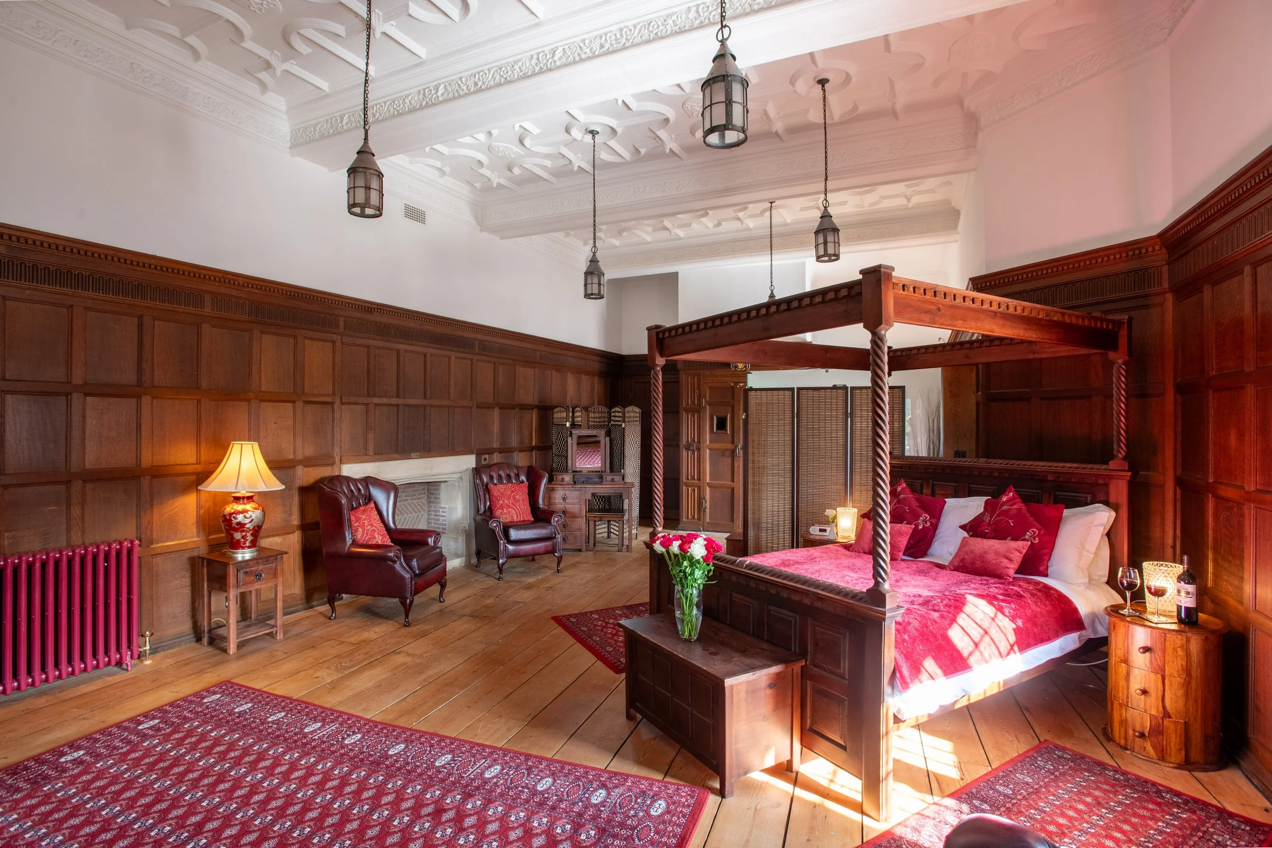 A luxurious bedroom with a four-poster bed, dark wooden paneling, leather chairs, a fireplace, and vintage-style lighting fixtures.