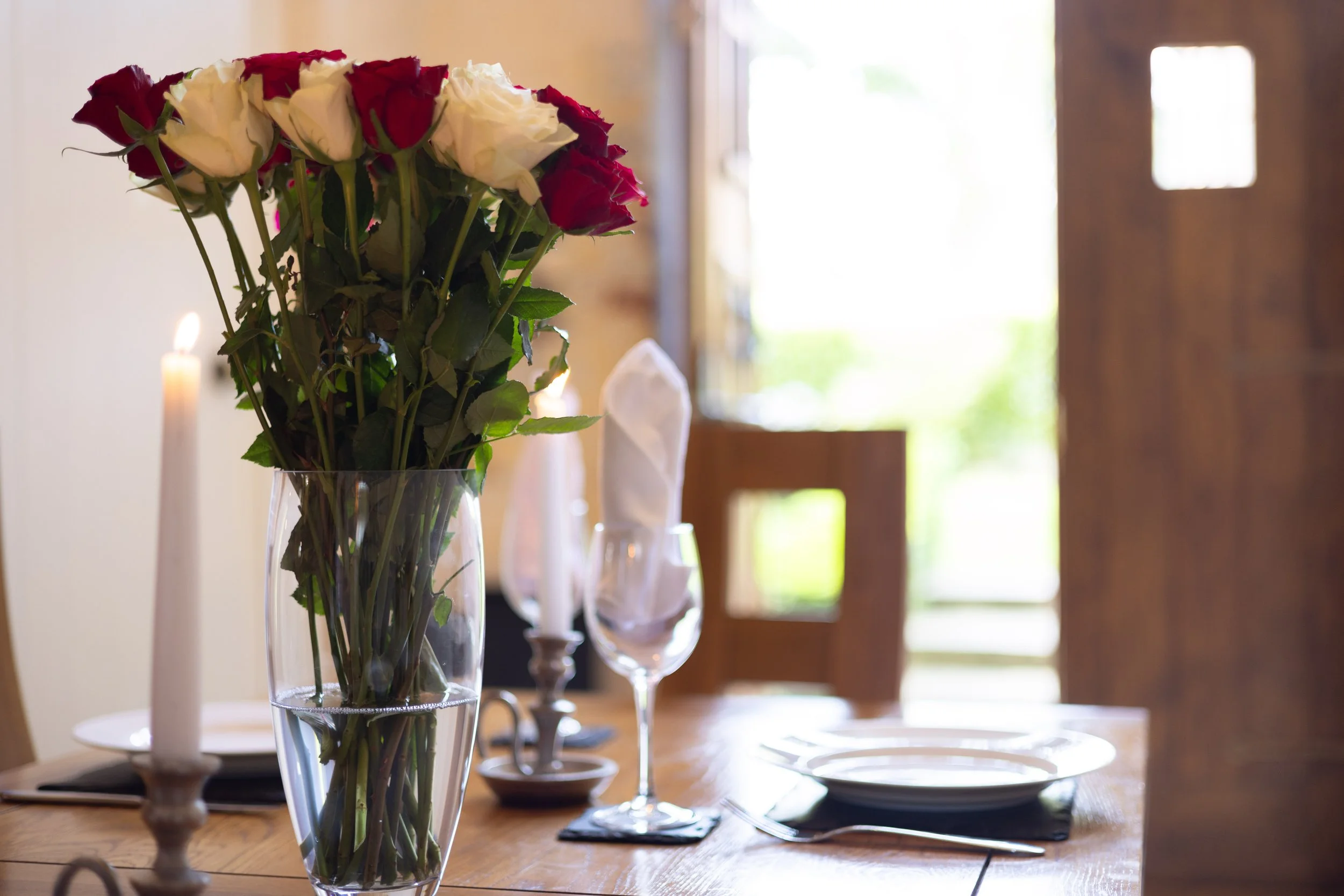 A dining table set with white plates, wine glasses, napkins, and candles, with a vase of red and white roses as the centerpiece, in a room with large windows.