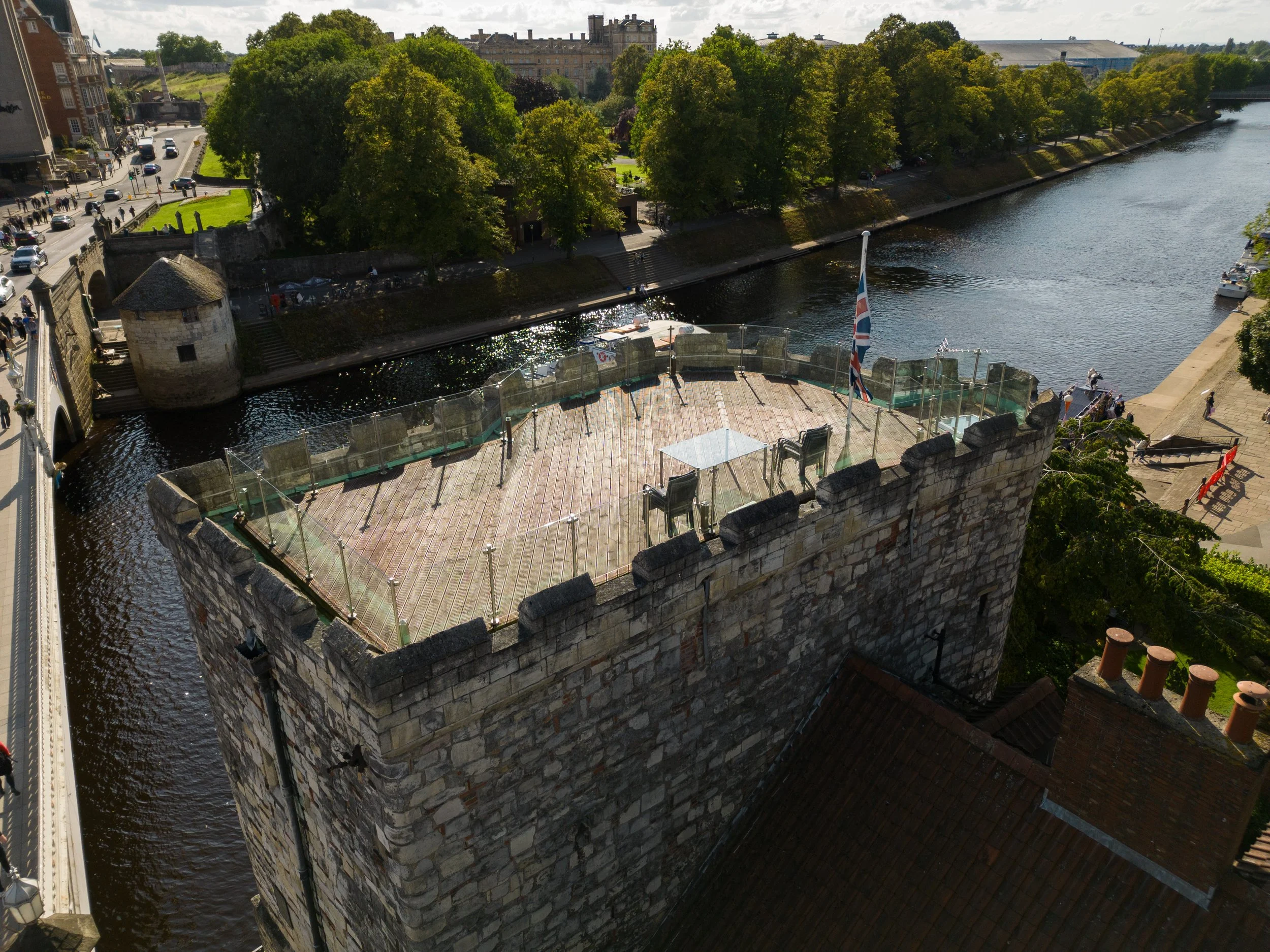 A historic stone tower with a rounded top situated on a corner of a castle wall overlooking a river. The wall has a wooden platform with chairs and an umbrella, bordered by a glass safety barrier. A Union Jack flag is seen near the platform. In the b