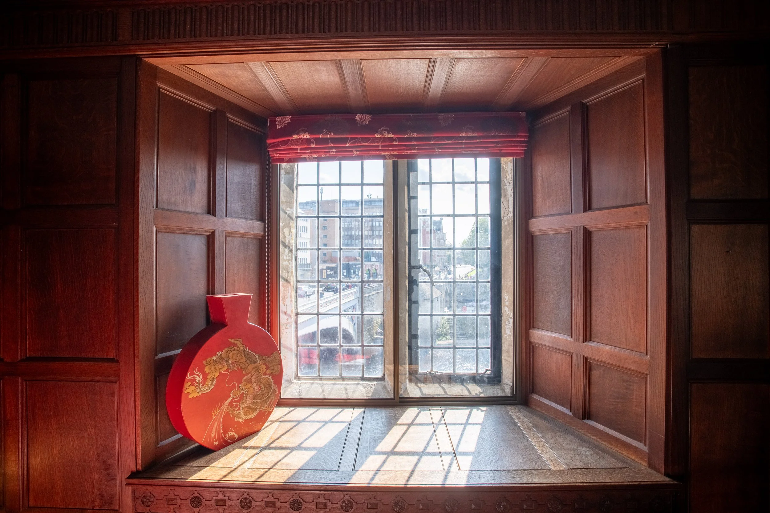 Wood-paneled window area with a red floral curtain valance and a view of city buildings outside. There is a red decorative object with a golden dragon design on the left side of the window.